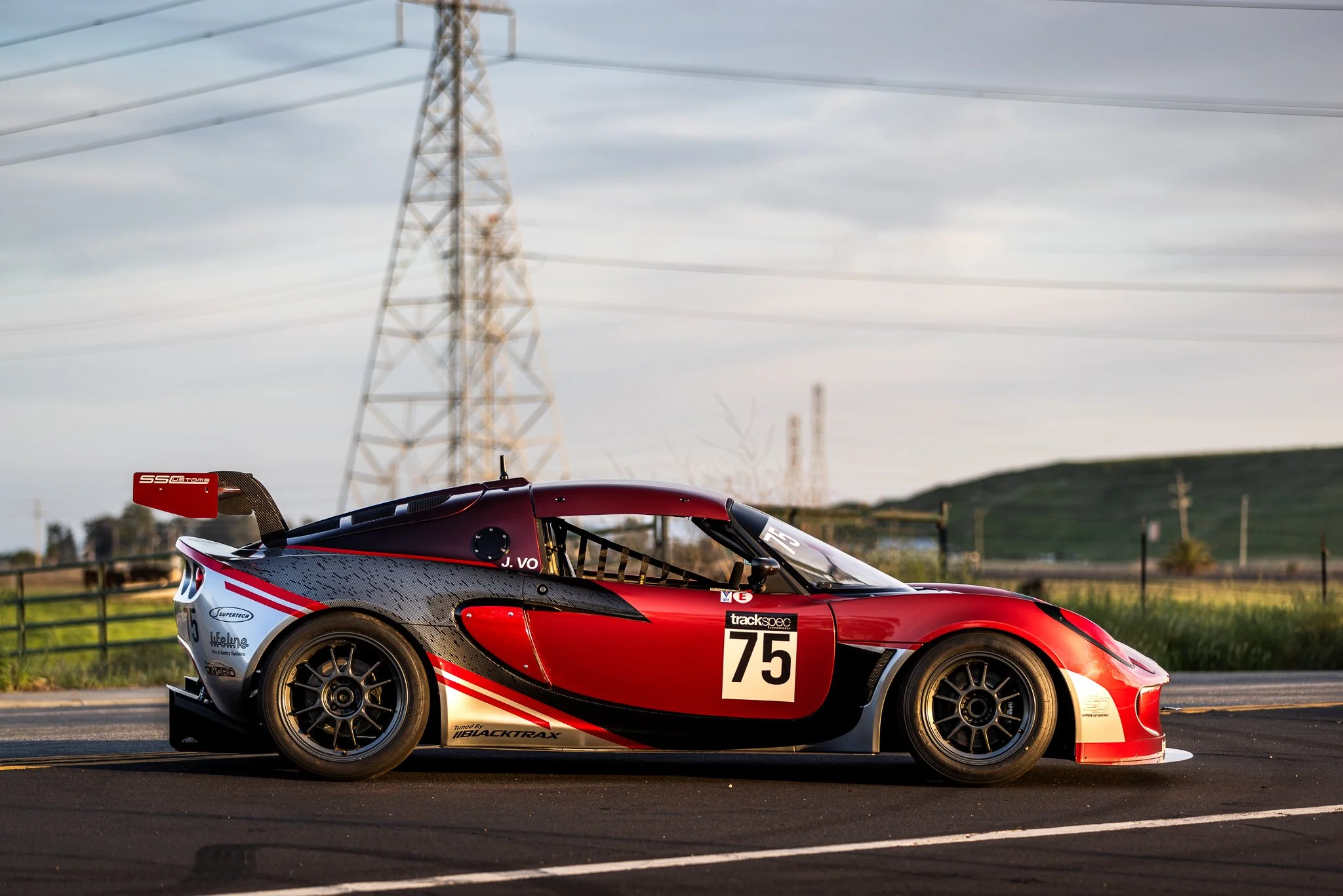 Red and black race car with the number 75 on the side parked on a track with grassy fields and power lines in the background.