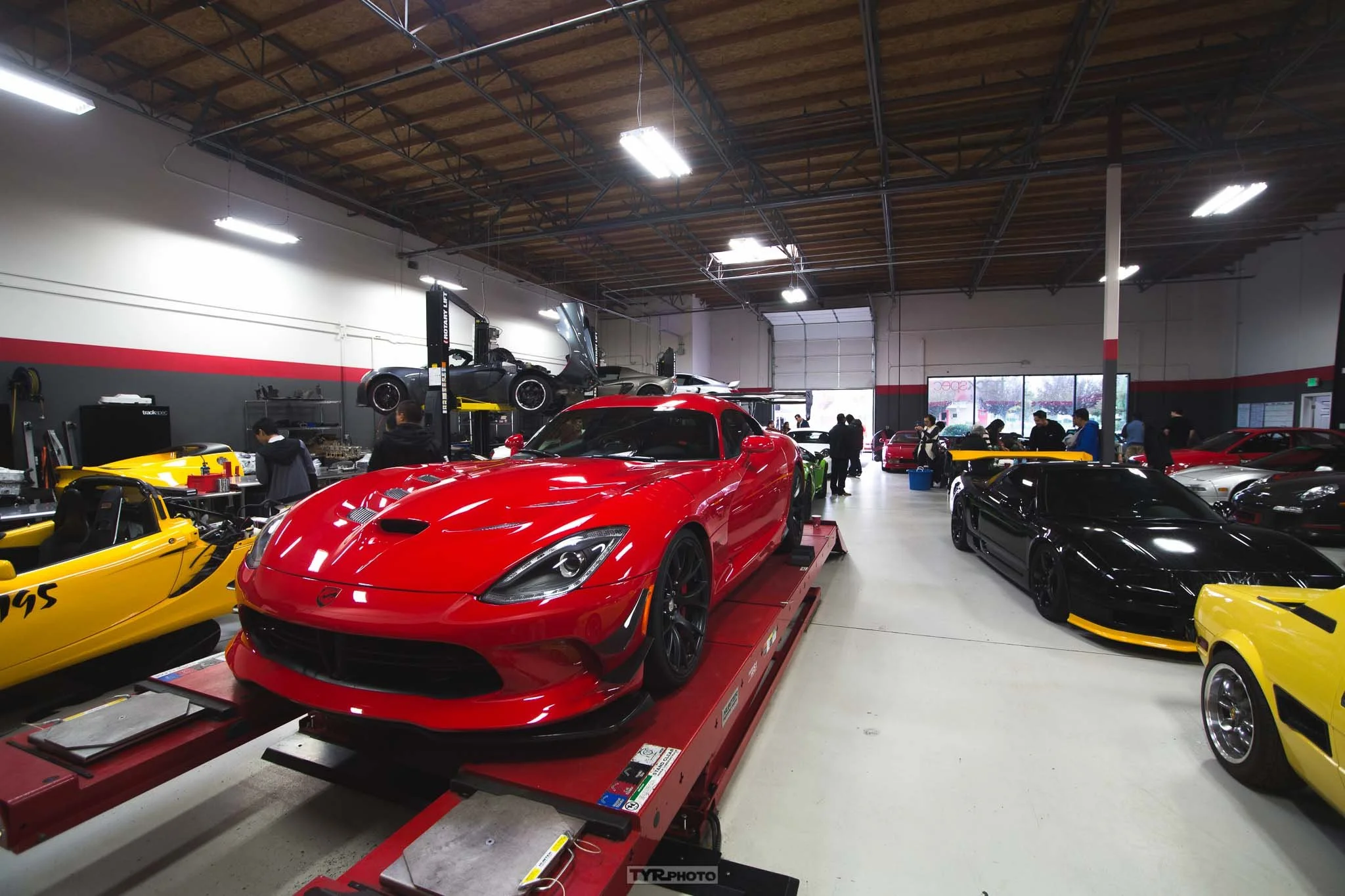 Interior of a car showroom with several high-performance cars on display, including a red sports car on a lift, a black car, a yellow race car, and a yellow vintage car; people inspecting and working on the vehicles.