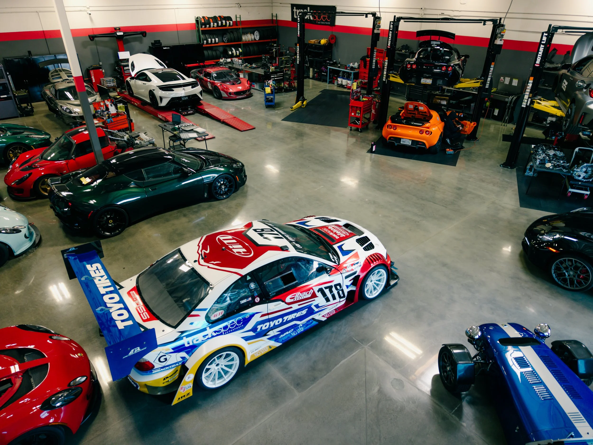 A spacious indoor garage filled with luxury and sports cars, including a white race car with sponsor logos, several high-end cars in various colors, and a car lift system in the background.