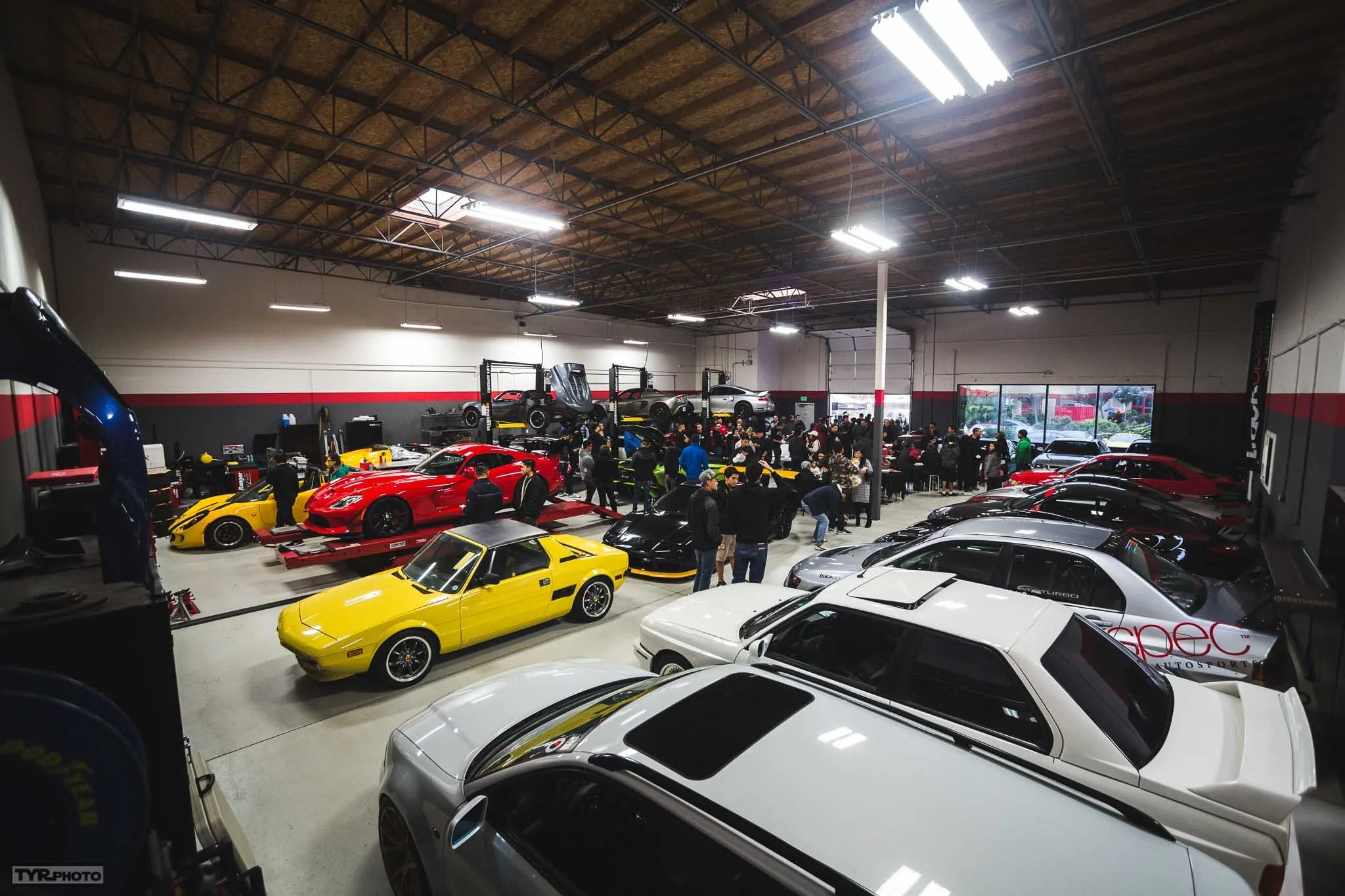 An indoor car showroom with a variety of sports cars including Ferrari, Lamborghini, and other performance vehicles, with many people inspecting and walking around the cars.
