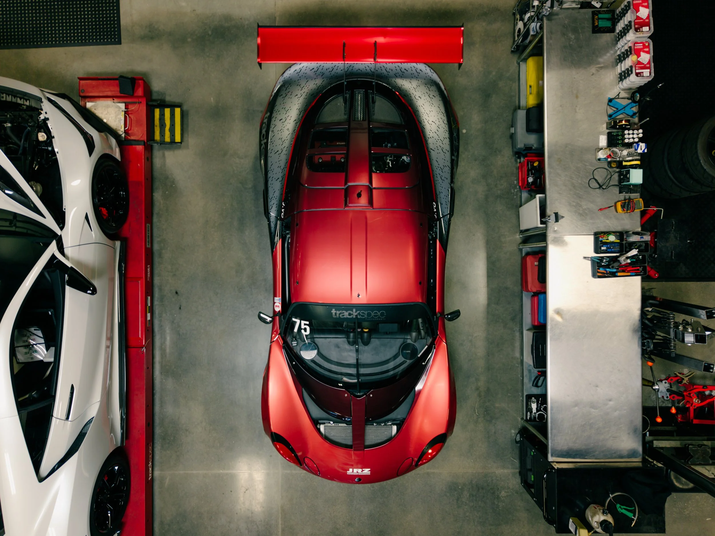 Top-down view of a red race car in a garage, with tools and equipment on workbenches around it, and a white car partially visible on the left.