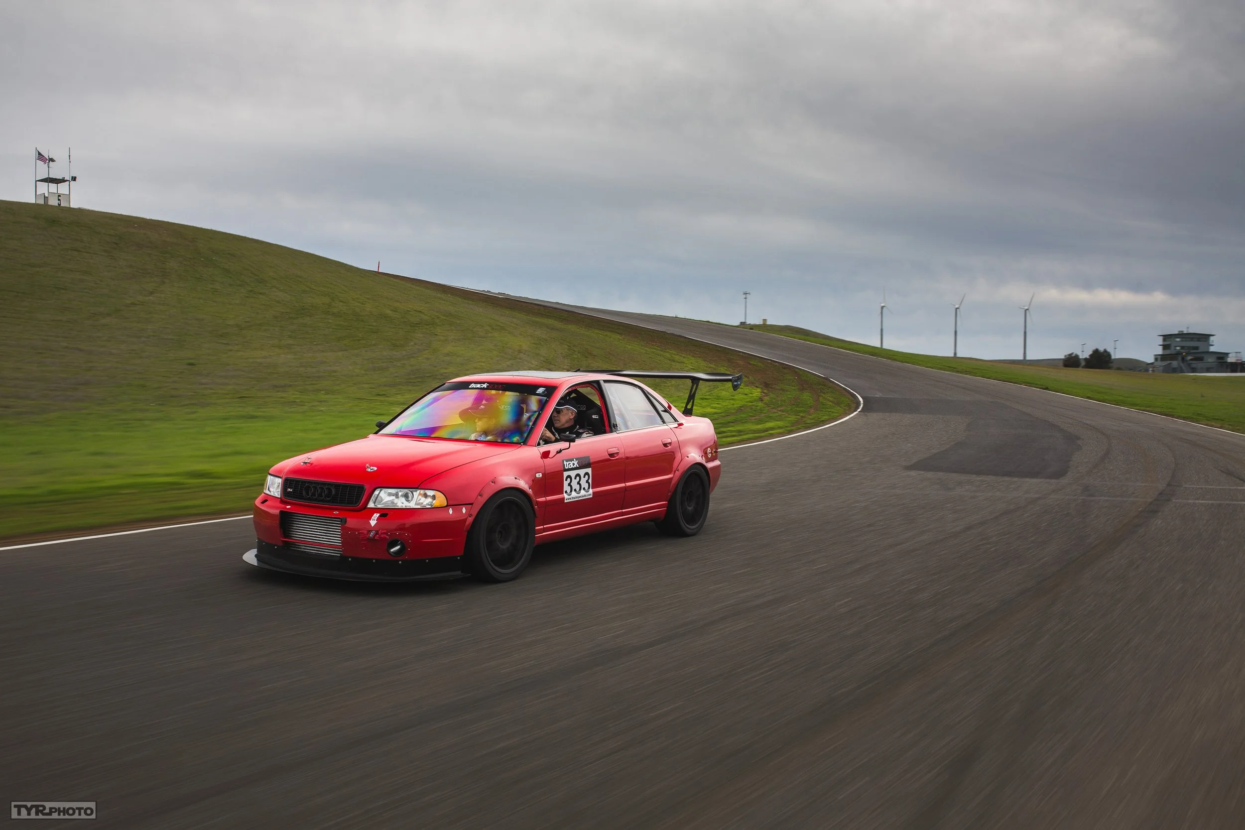 Red race car with the number 333 on the side driving on a curved race track under cloudy sky.