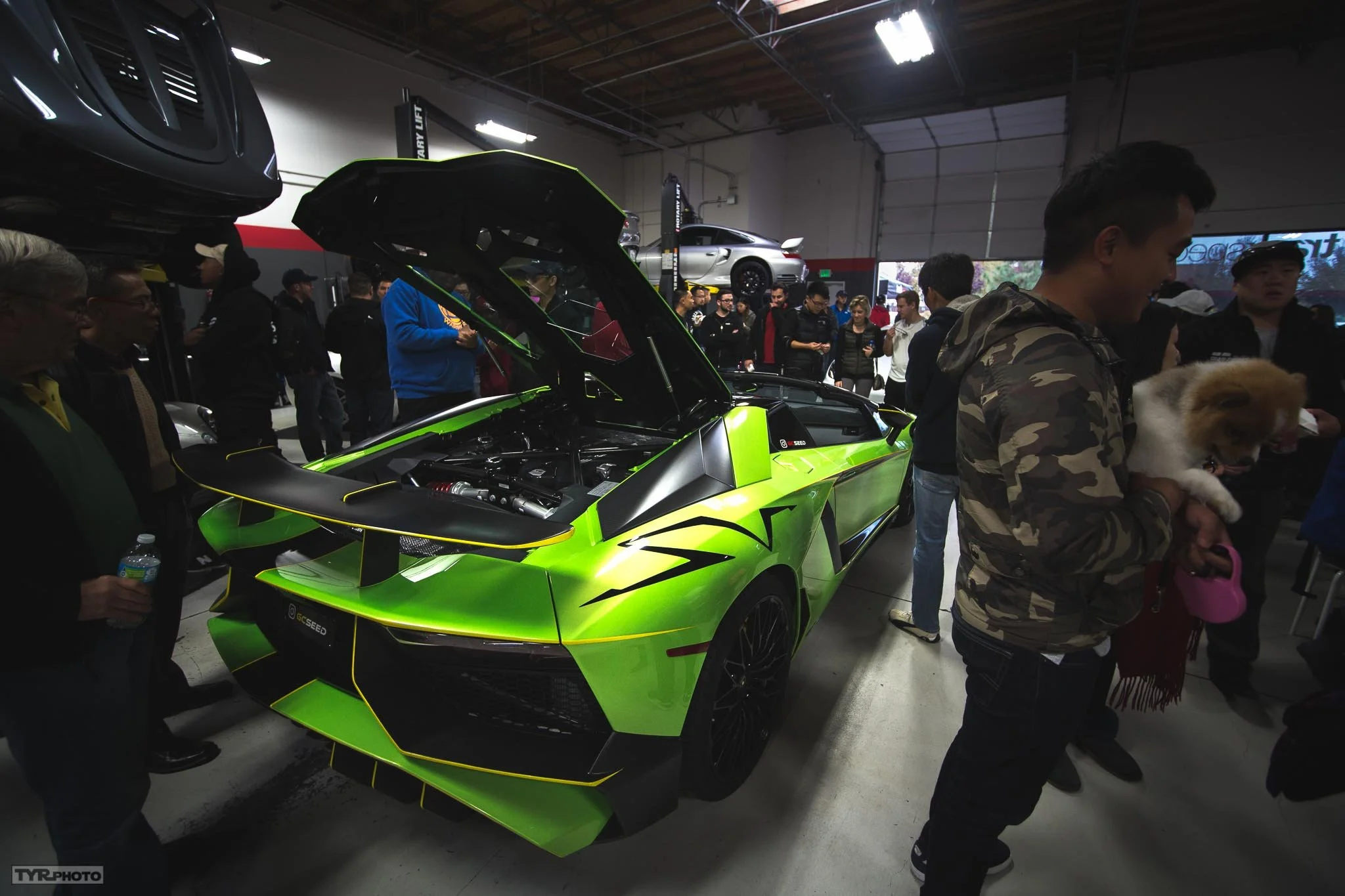 Green Lamborghini sports car with its hood open on display at an auto show, surrounded by people inside a large industrial-style building.