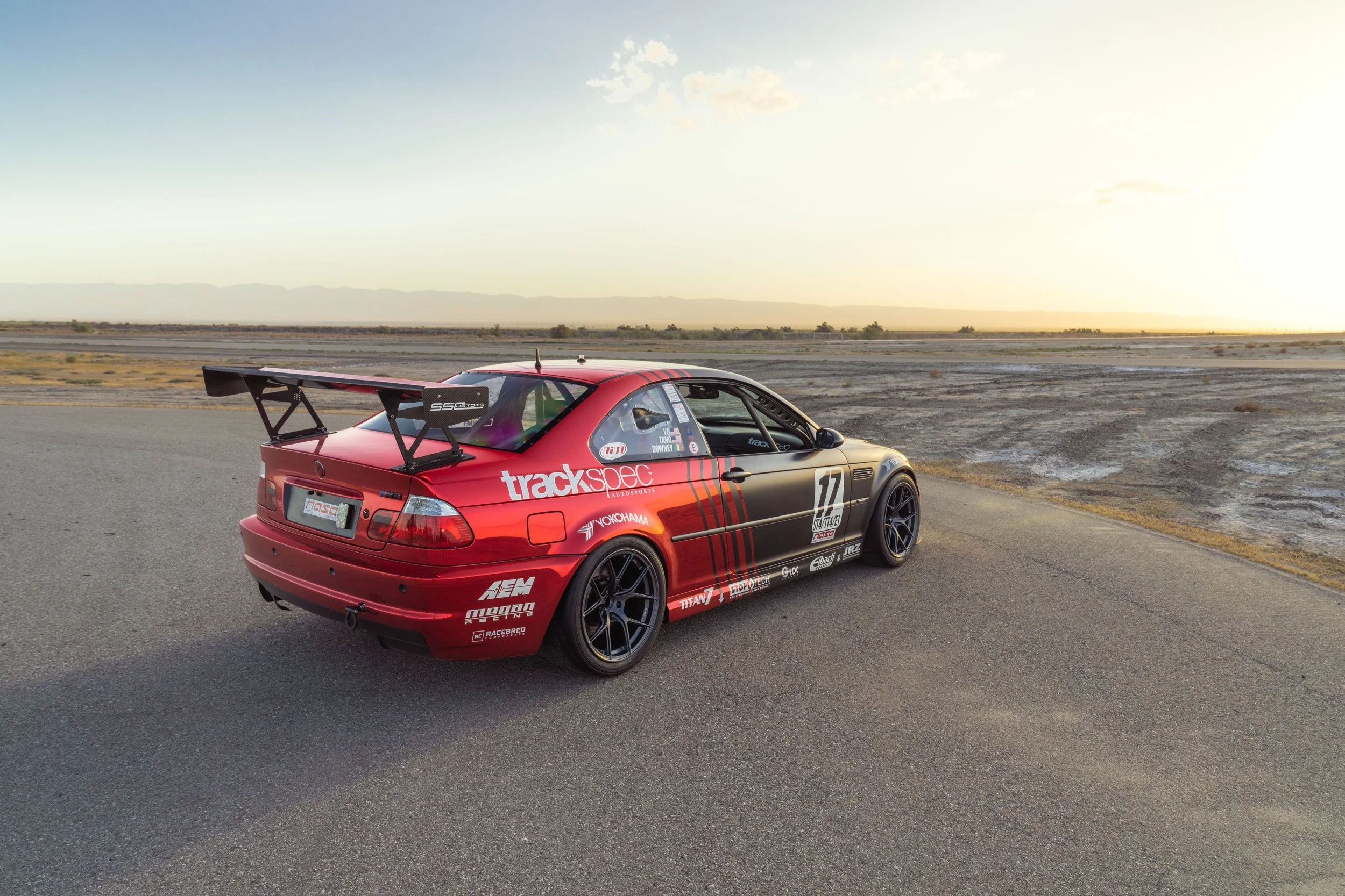 Red race car with black accents, a large rear wing, and sponsorship decals, parked on the side of a deserted road at sunset.