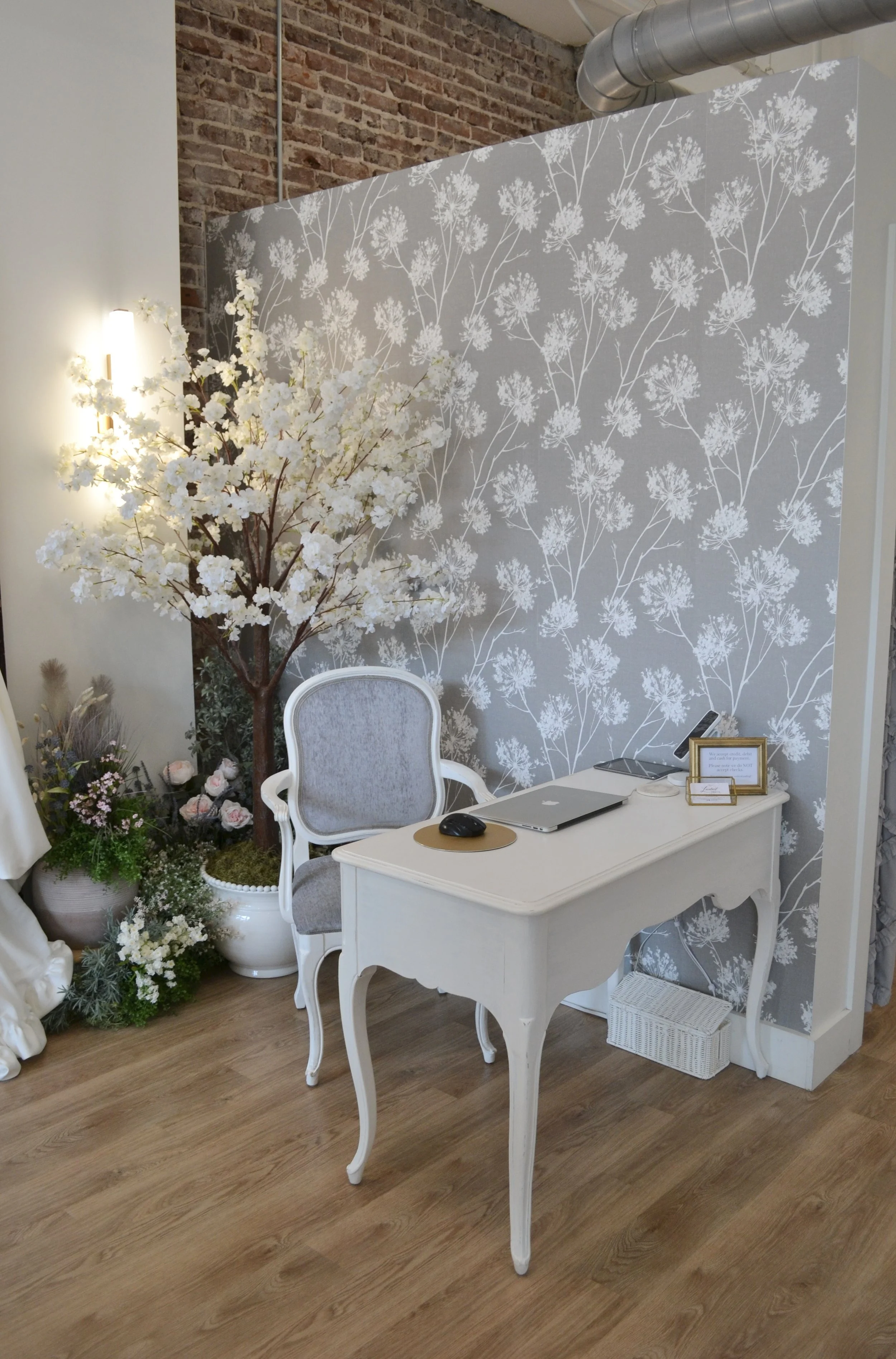 A white vintage-style desk with a laptop, mouse, and framed sign, accompanied by a white upholstered chair, in a room with floral wallpaper, a faux cherry blossom tree, and floral arrangements.