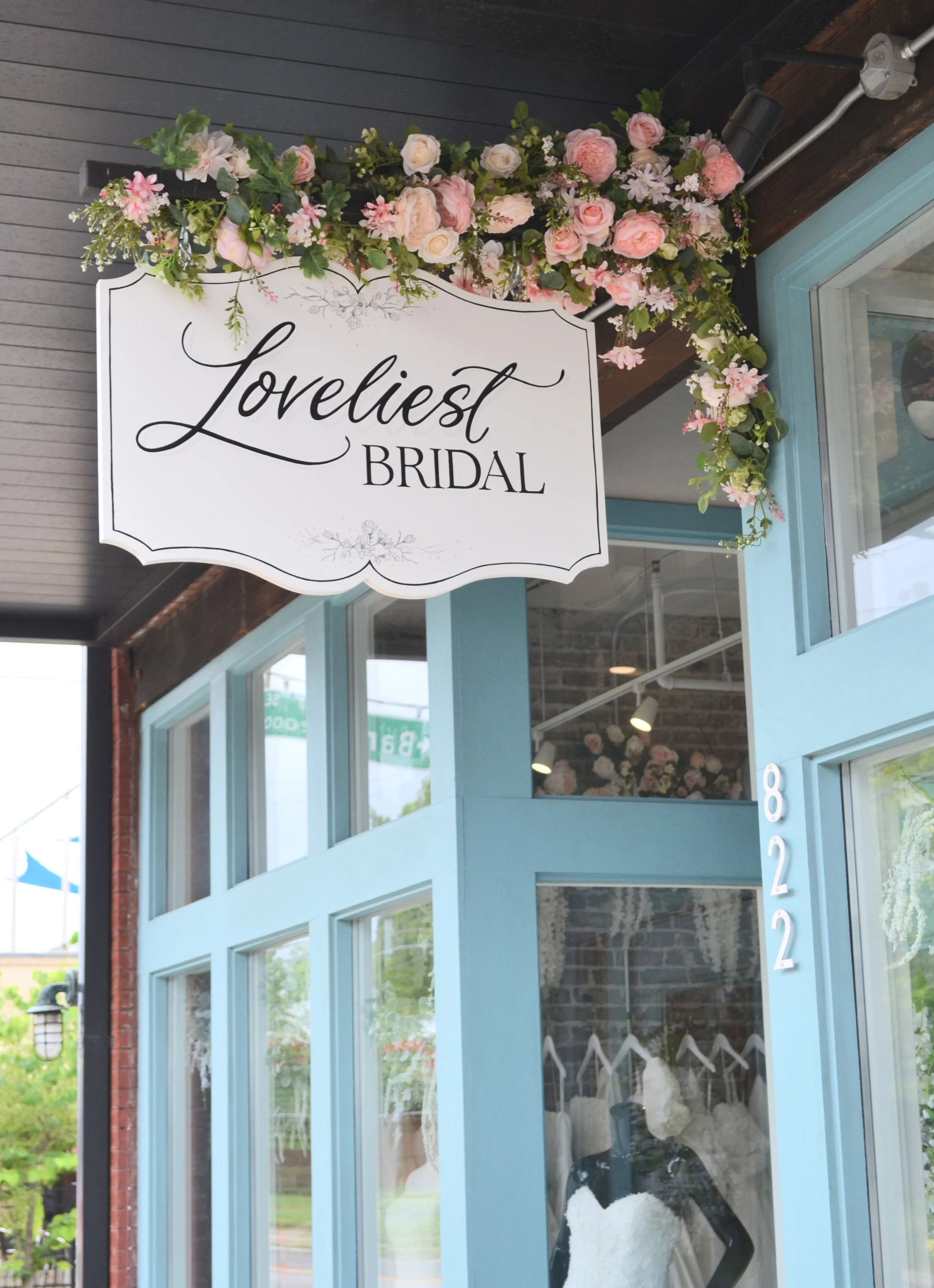 The exterior of a bridal boutique decorated with a sign that reads 'Loveliest Bridal' and a floral arrangement of pink and white flowers above the entrance. The building has large glass windows and a light blue door with the number 8622.