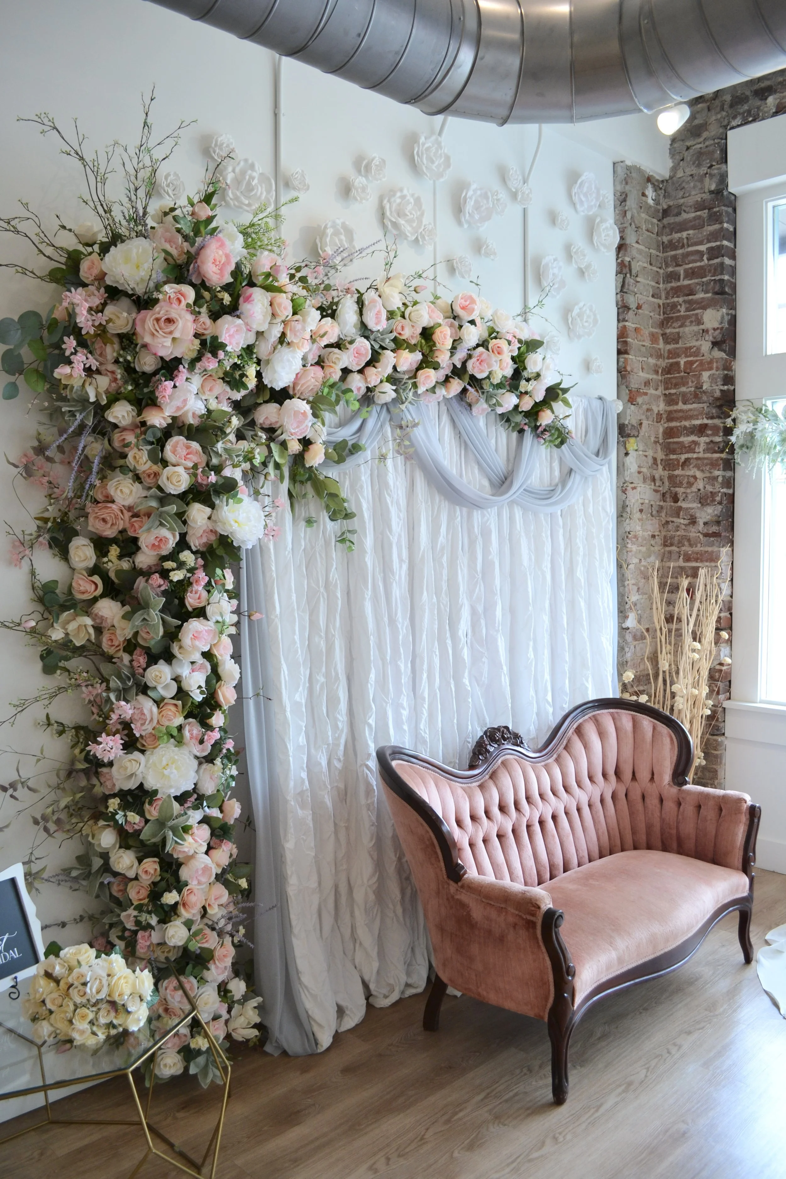 A floral wedding backdrop with pink and white roses, green foliage, and branches, decorated with white fabric drapes and hanging paper flowers. There is a vintage pink velvet loveseat and a gold geometric side table with more white roses in the room with exposed brick and white walls.