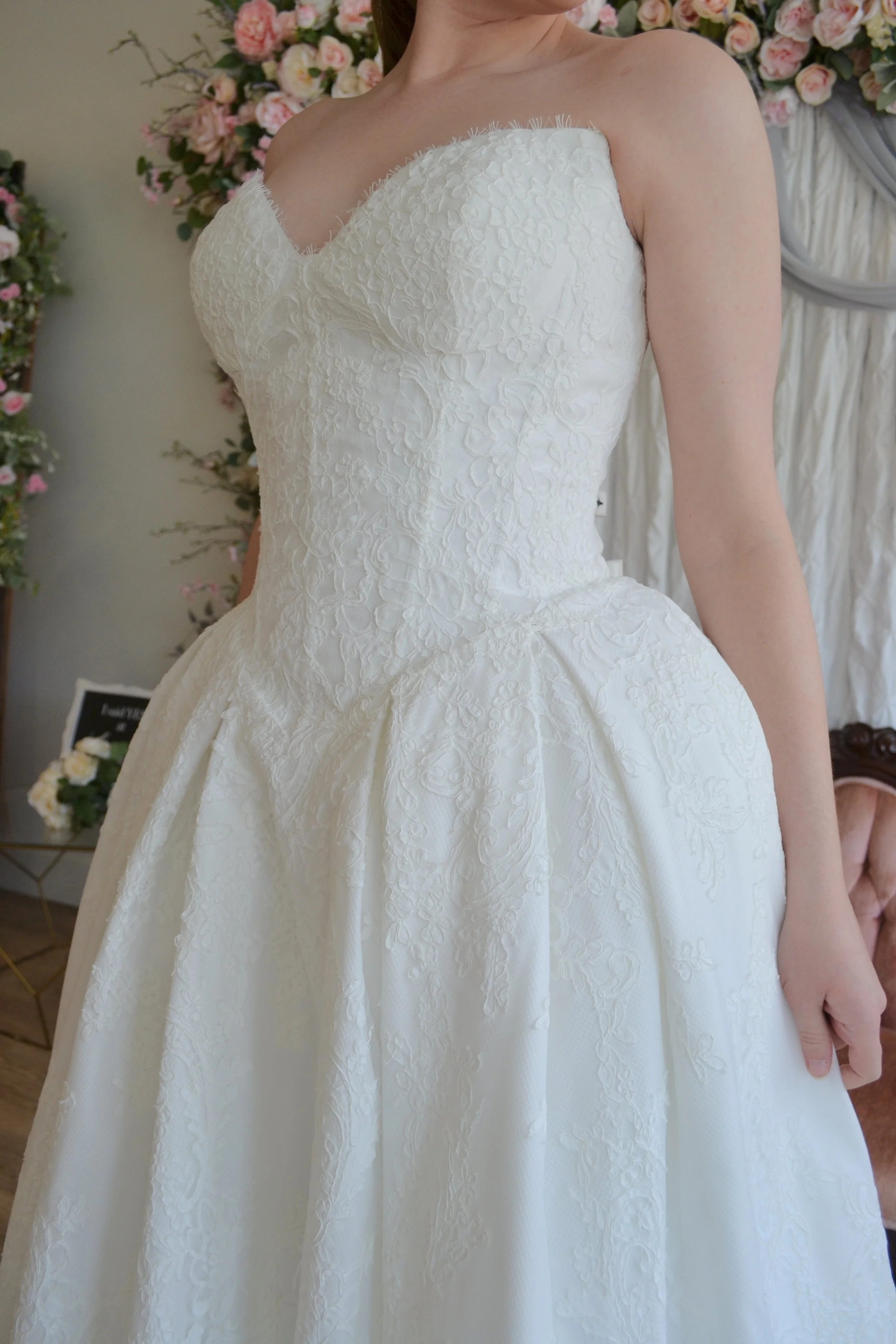 Close-up of a woman in a wedding dress with a fitted bodice and a full skirt, standing in front of a floral background of pink and white roses.