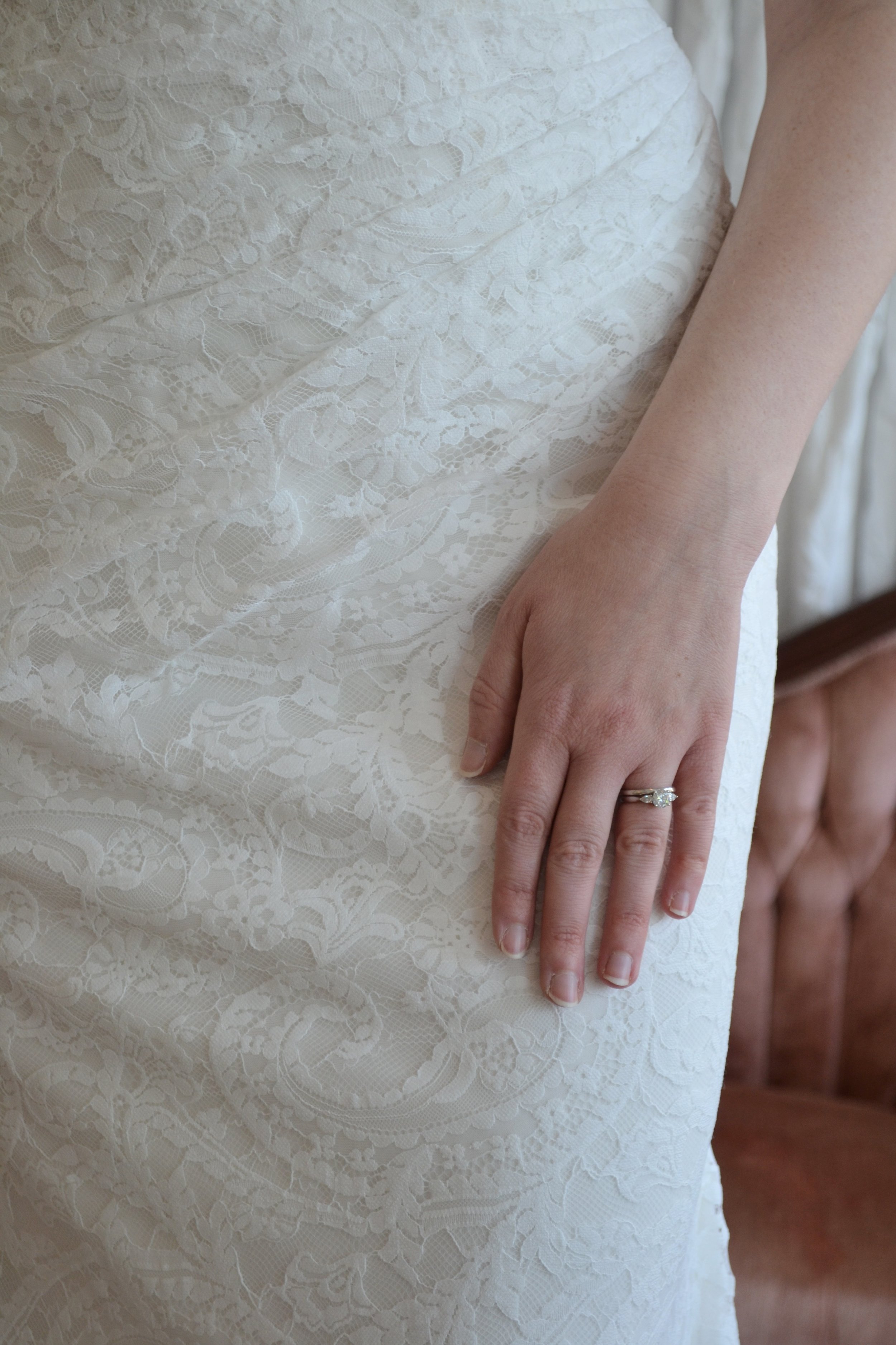 Close-up of a woman's hand resting on her waist, wearing a marriage ring, next to a white lace wedding dress.