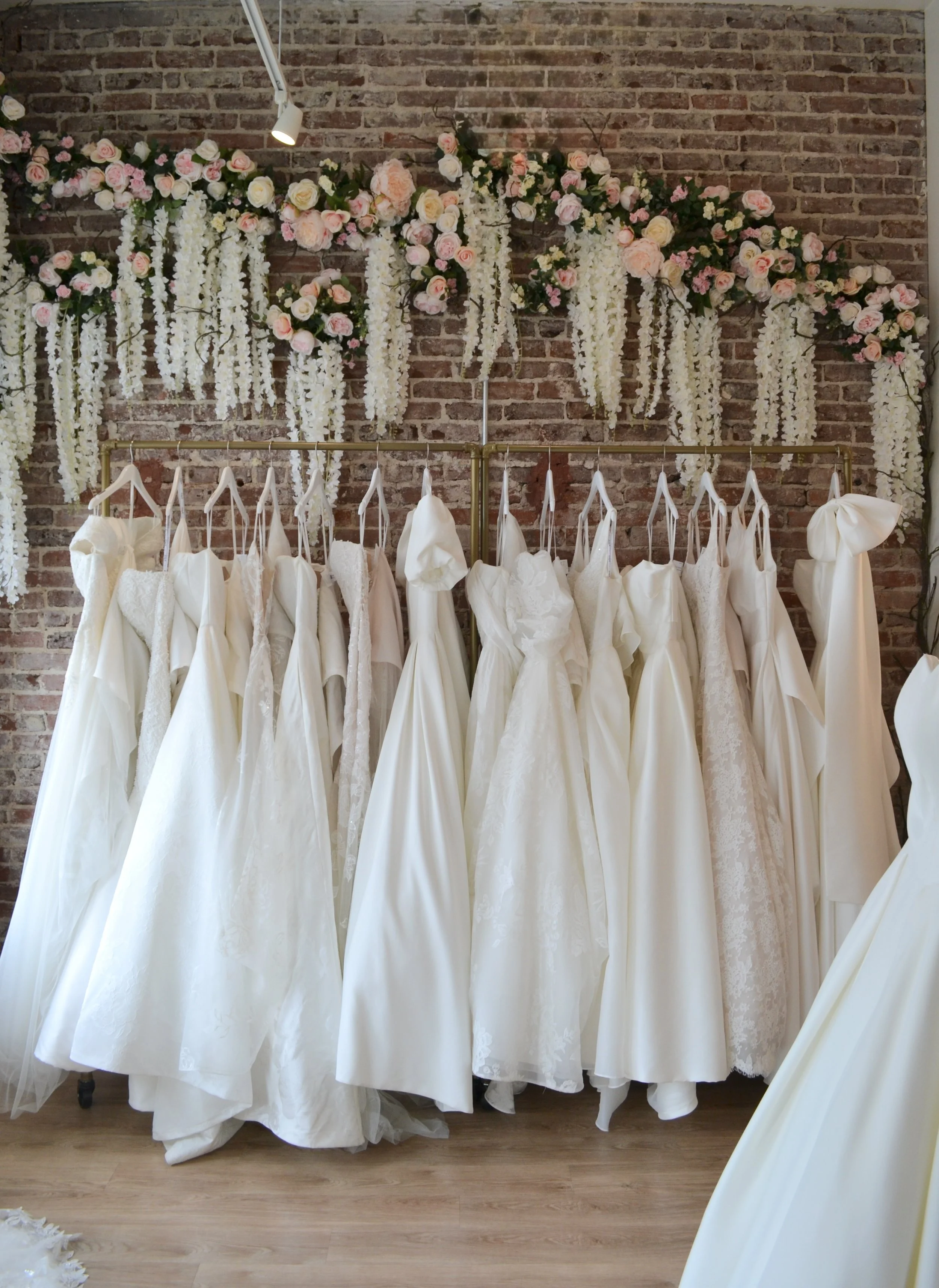 A display of wedding dresses hanging on a clothing rack in front of a brick wall arch decorated with white and pink flowers and hanging white floral garlands.