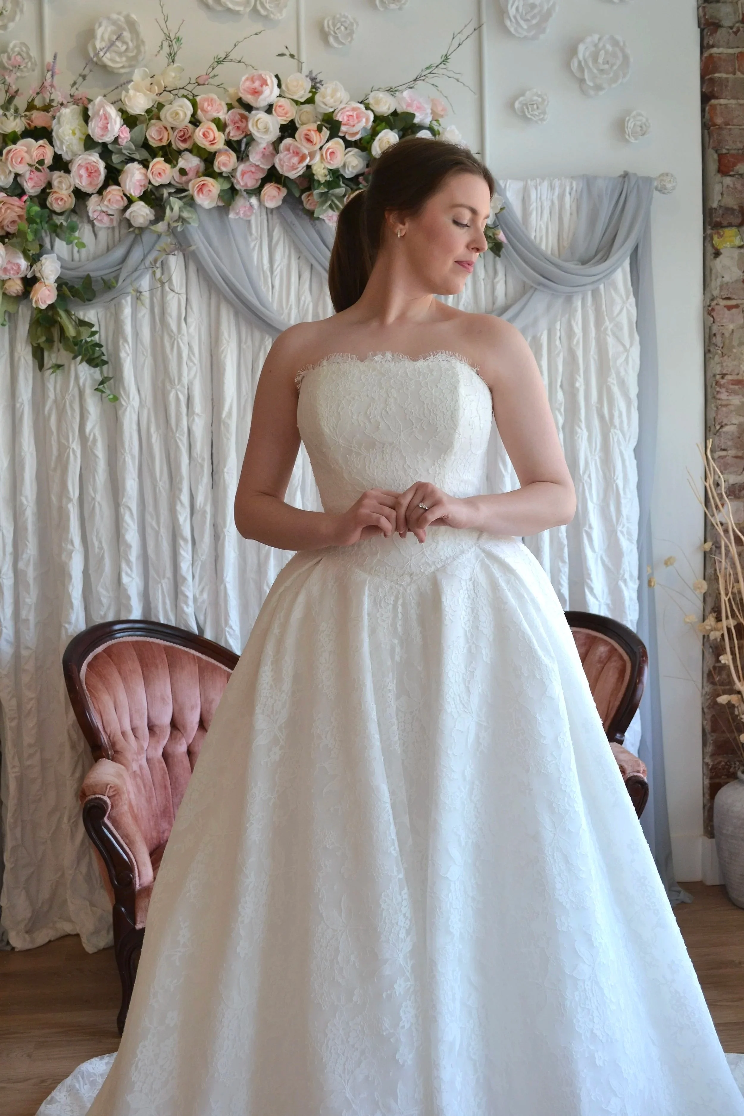 A bride in a white lace strapless wedding dress, standing with her eyes closed and hands clasped in front of her, in a decorated room with a floral backdrop and velvet chairs.