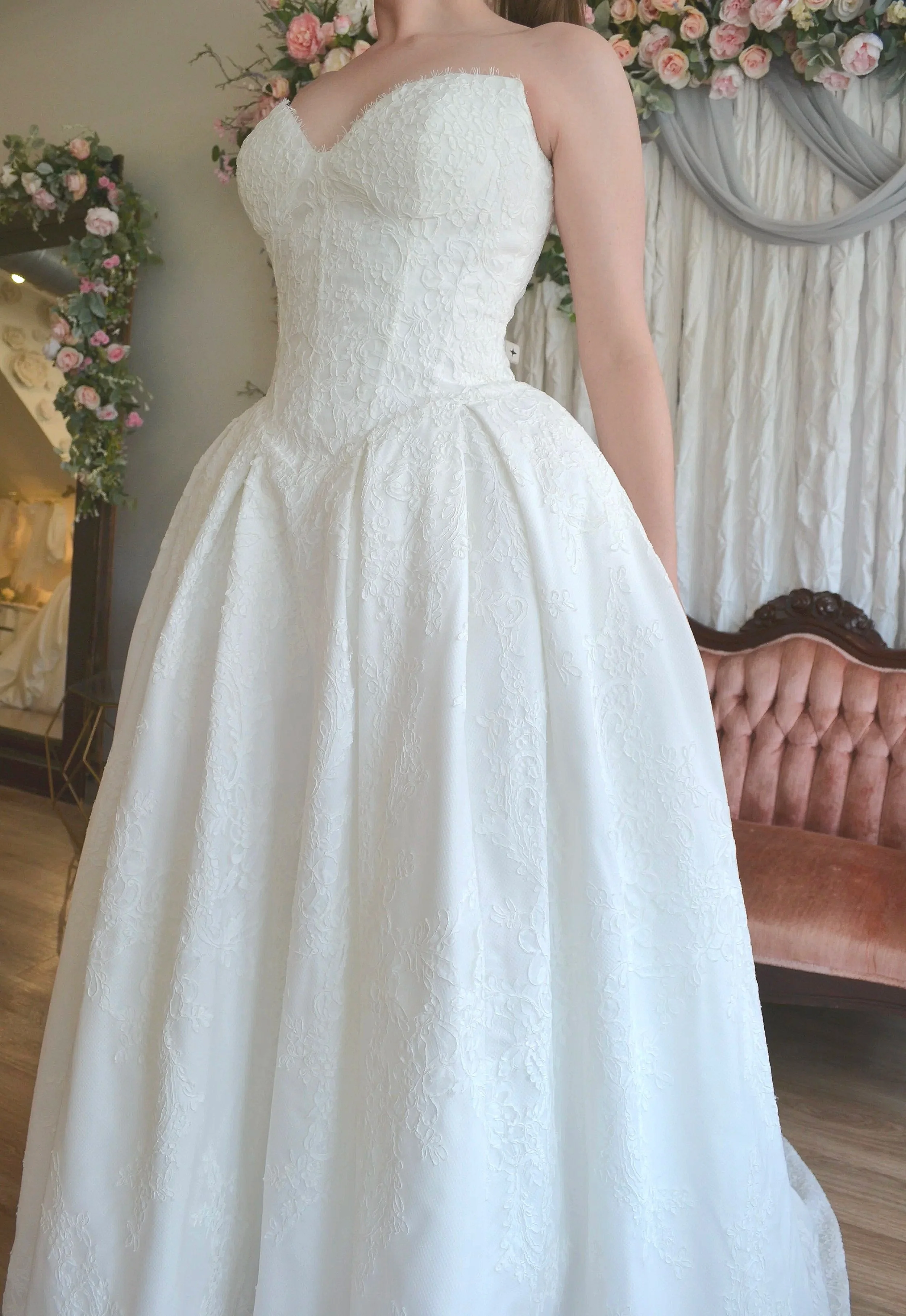 A bride in a wedding dress standing indoors, with pastel pink roses and greenery in the background, and a velvet armchair partially visible.