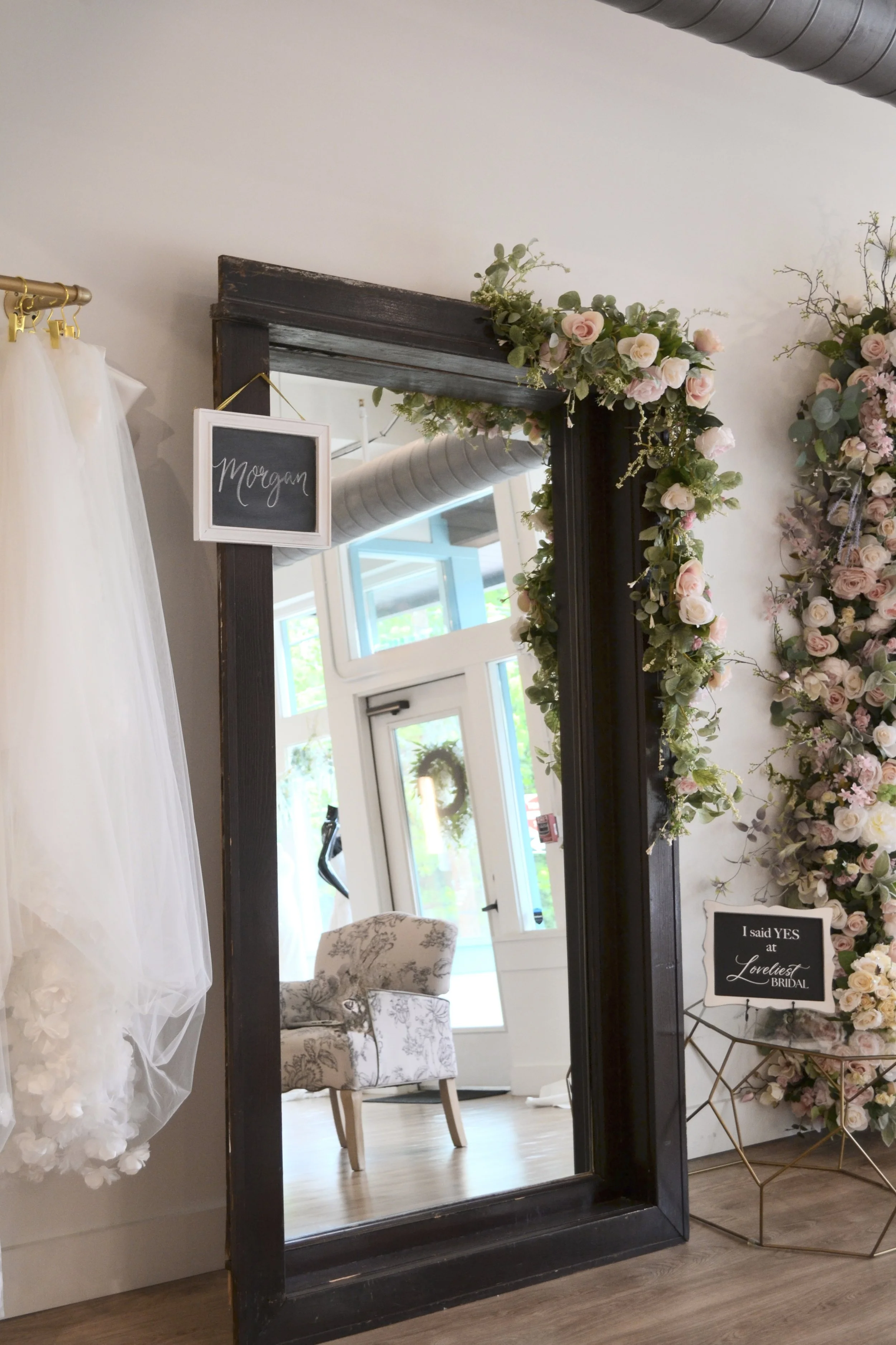 A mirror decorated with pink and white roses and green foliage, with black frames. Reflects a white wall with a window and a beige floral chair, and part of a white door. To the left, a white curtain with gold hooks. To the right, a gold side table with a black sign reading 'I said YES at Loveliest Bridal'.