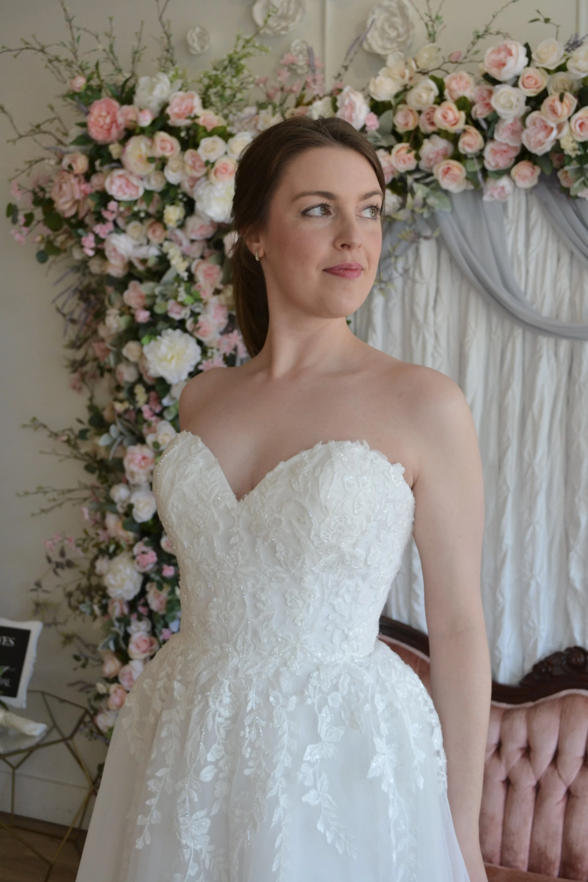 A woman in a white wedding dress sitting in front of a large floral arrangement with pink and white roses and greenery, inside a decorated room.