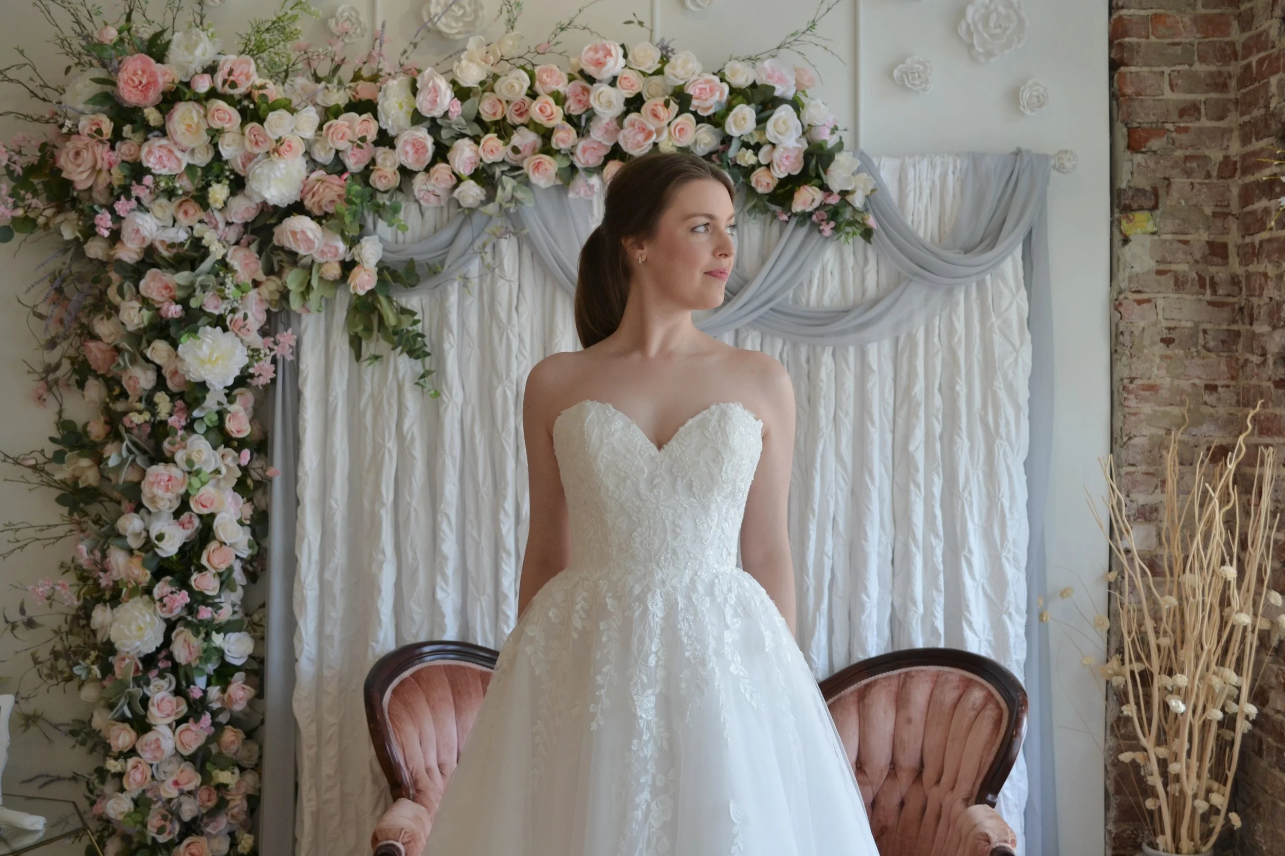 A woman in a wedding dress standing in front of a floral backdrop with pink and white roses and greenery.