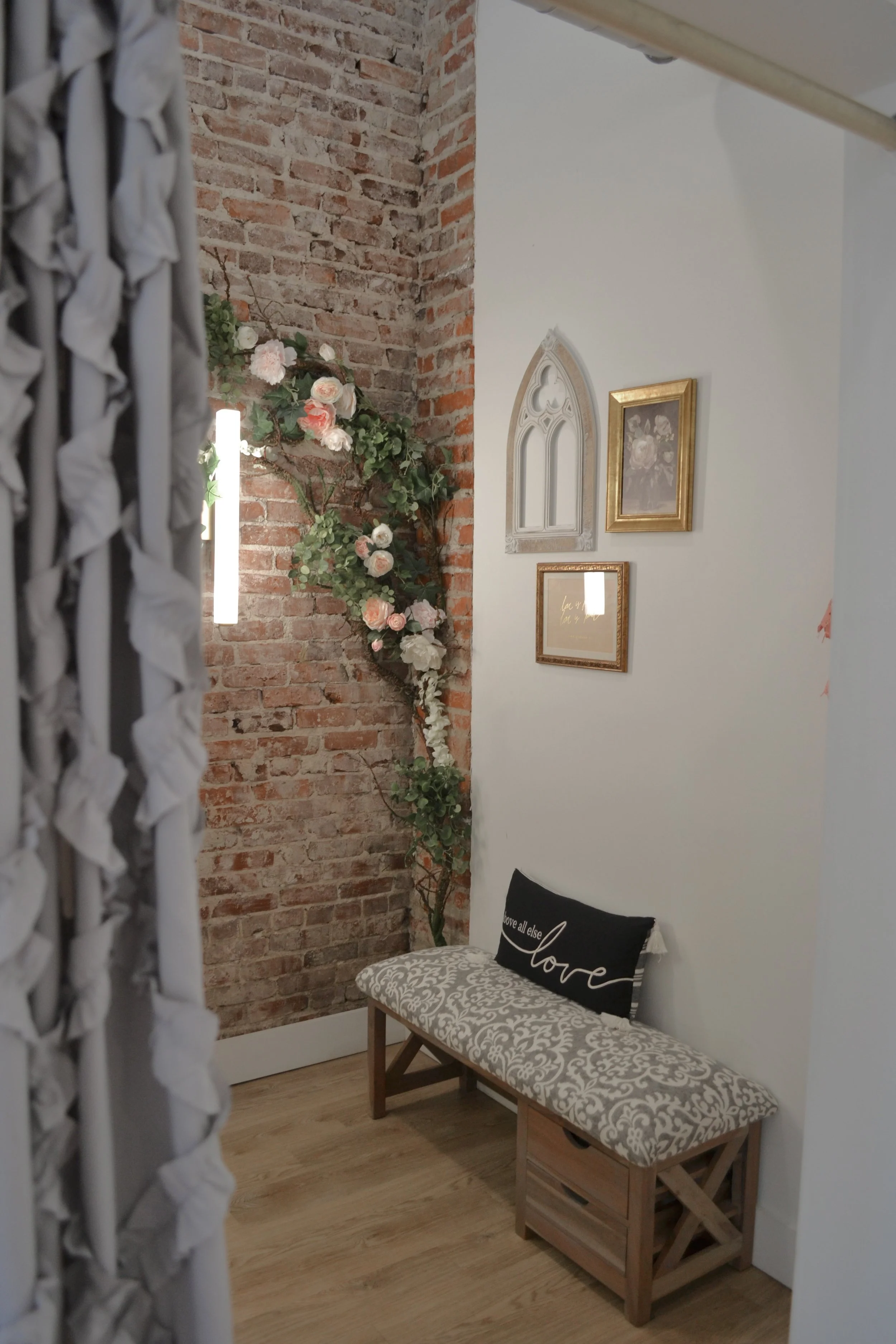 A cozy corner with a brick wall, a bench with a decorative pillow that says 'love,' floral wall decor, framed artwork, and a white curtain on the side.