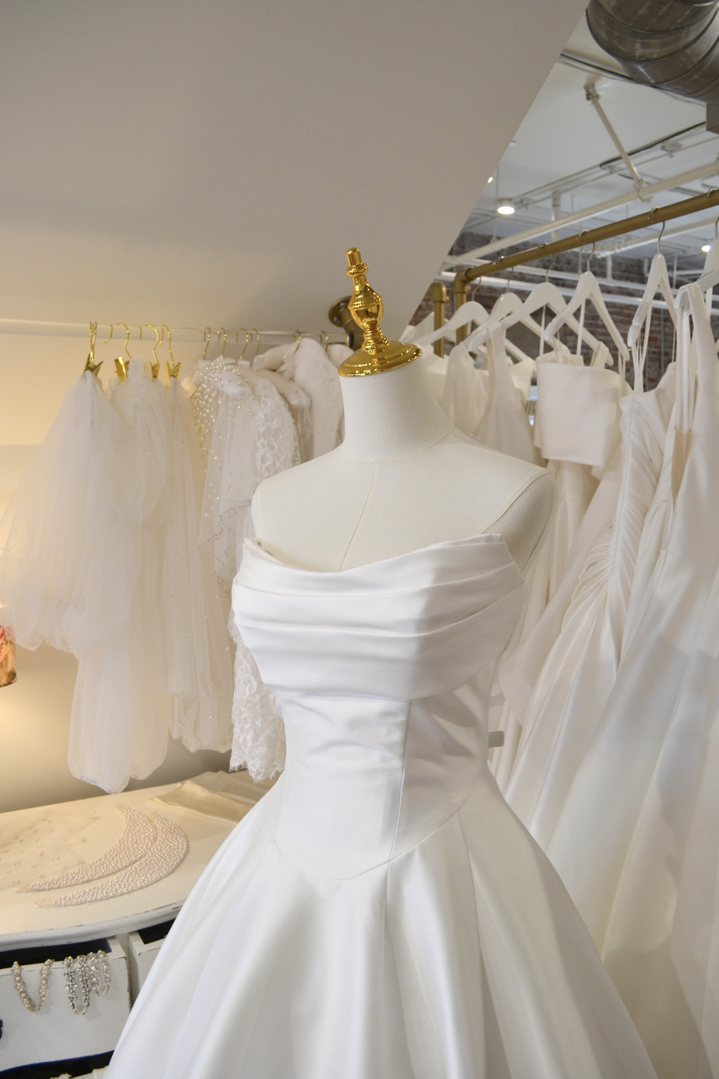 A mannequin dressed in a white wedding gown, surrounded by other wedding dresses and accessories in a bridal boutique.