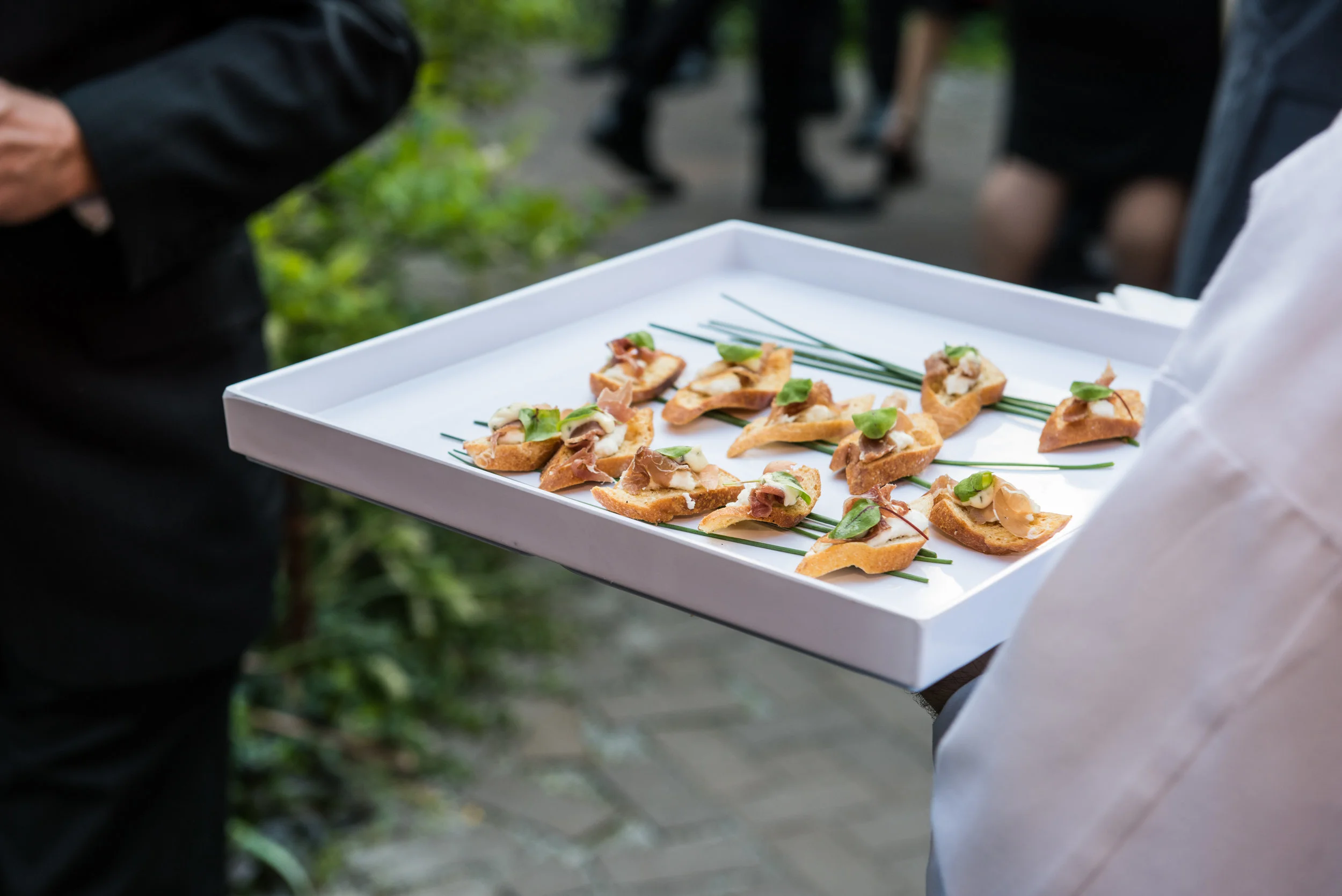 A server presenting a white tray of individually garnished hors d’oeuvres at an outdoor reception.