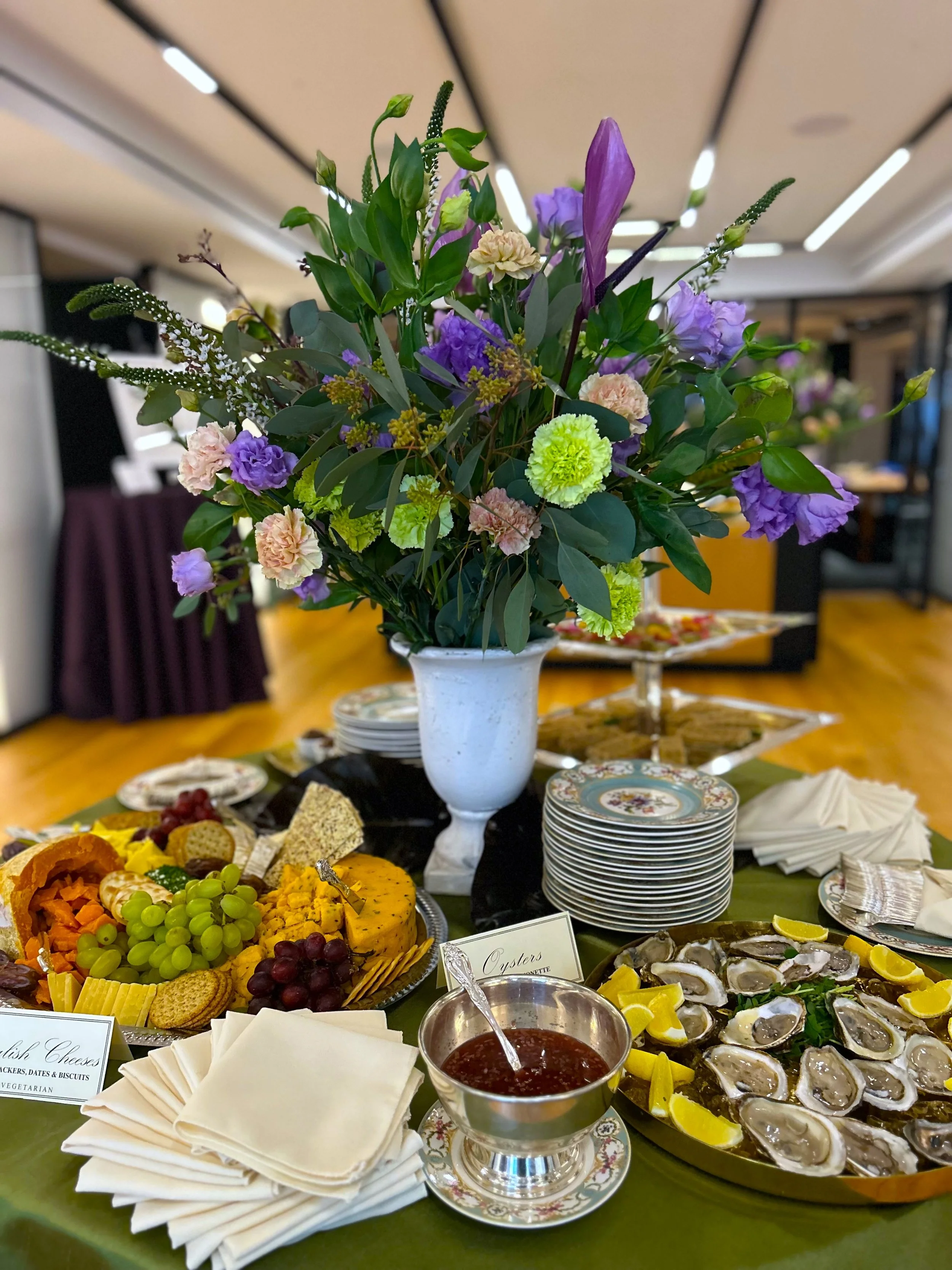 A reception display with oysters on ice, cheeses, grapes, stacked plates and a tall arrangement of purple and green flowers.