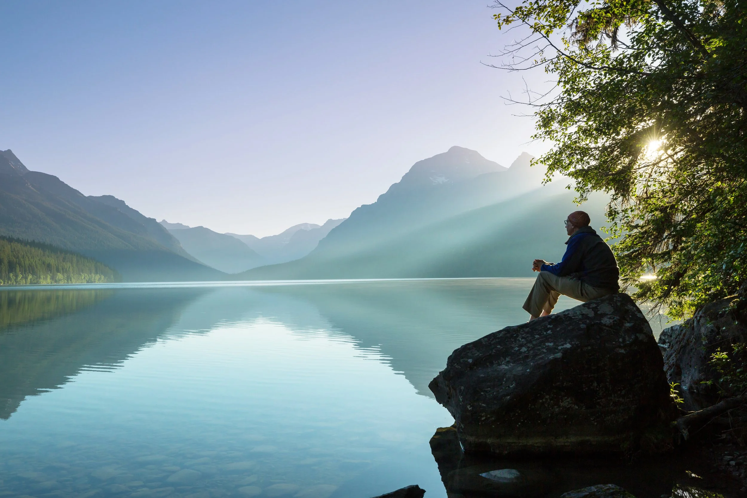 Man looking across a peaceful lake with mountains in the distance.