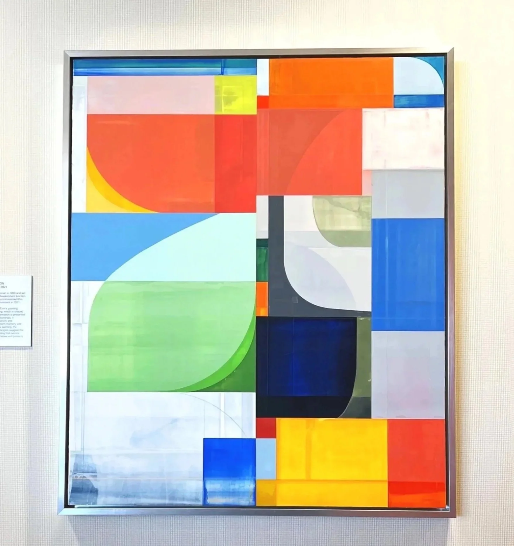 Abstract geometric artwork featuring colorful blocks, circles, and rectangles in red, yellow, blue, green, white, black, and orange tones on a gallery wall.