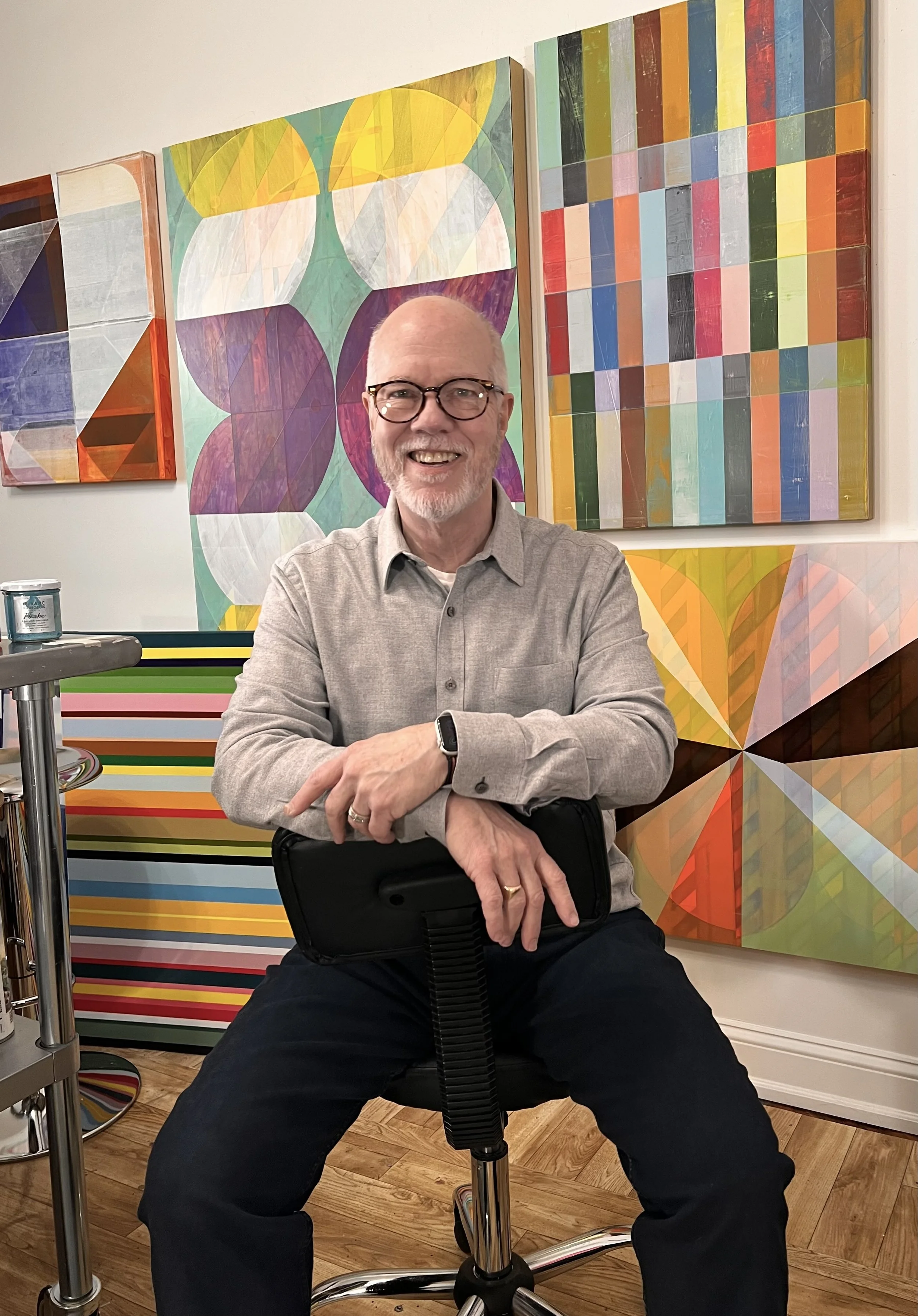 A smiling artist with glasses and a gray shirt sitting on a black chair in front of colorful abstract paintings in an art gallery or studio.