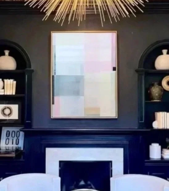 Living room with dark wall, white fireplace, built-in shelves with books and vases, and a large abstract painting. Modern chandelier hangs from the ceiling.
