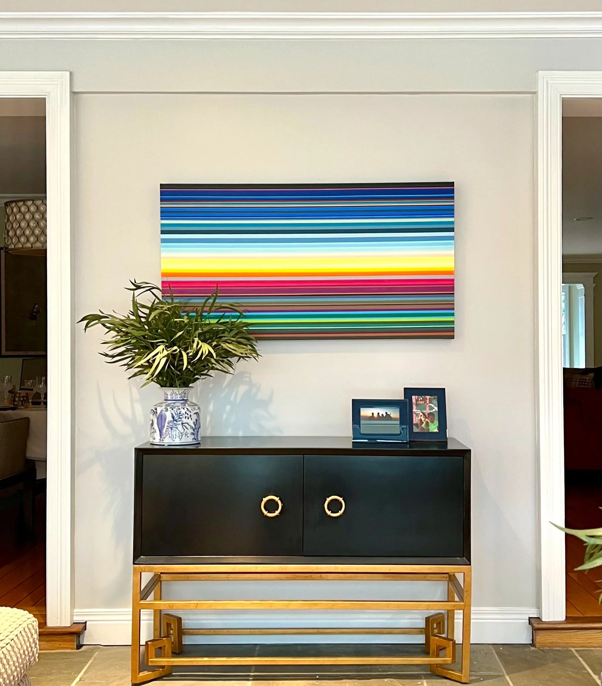A long horizontal striped painting in the colors of the sunrise hanging on an off white wall above a Japanese-syle credenza