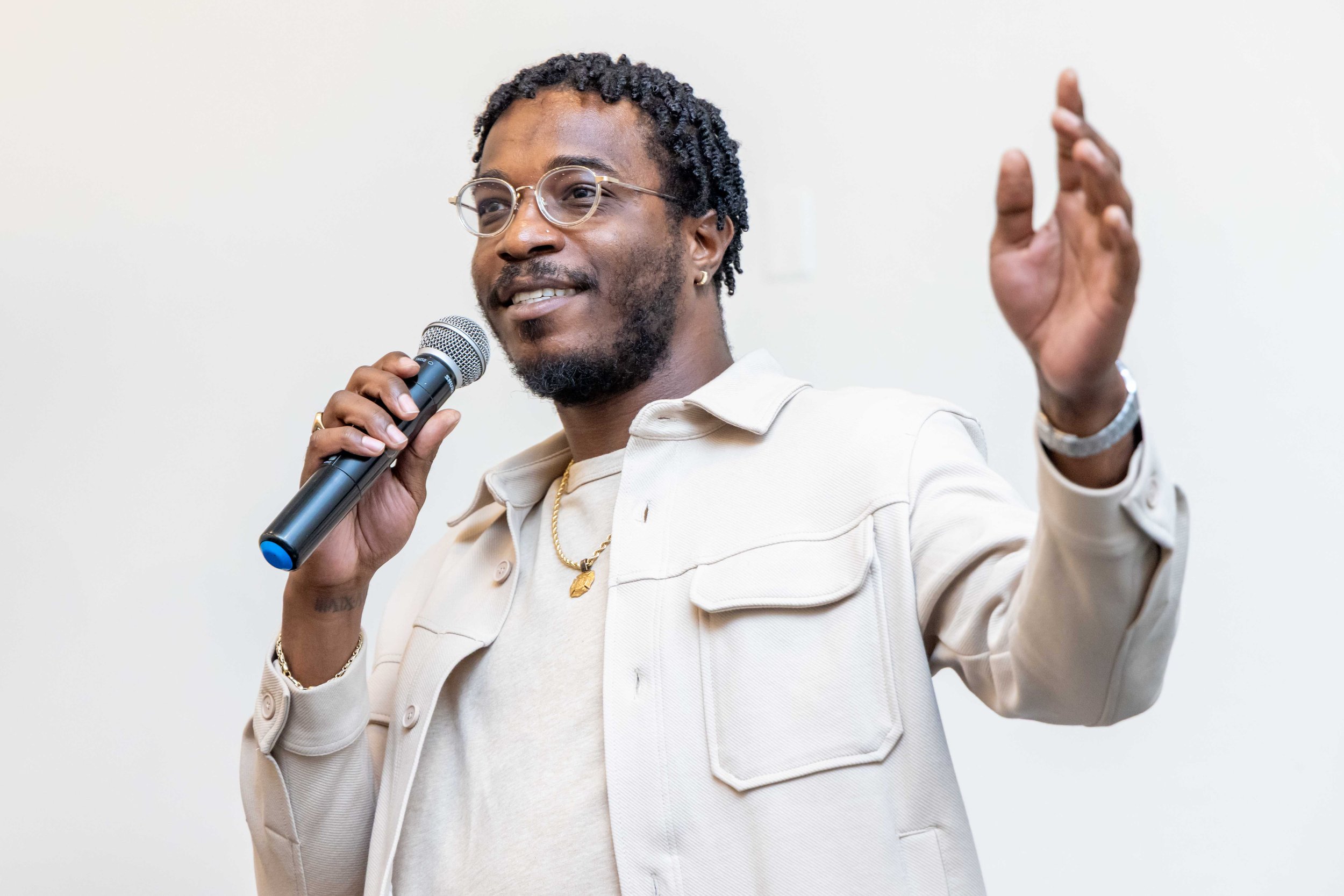 A man with glasses and a beard holding a microphone and speaking, wearing a beige jacket and white shirt, with his hand raised against a plain white background.