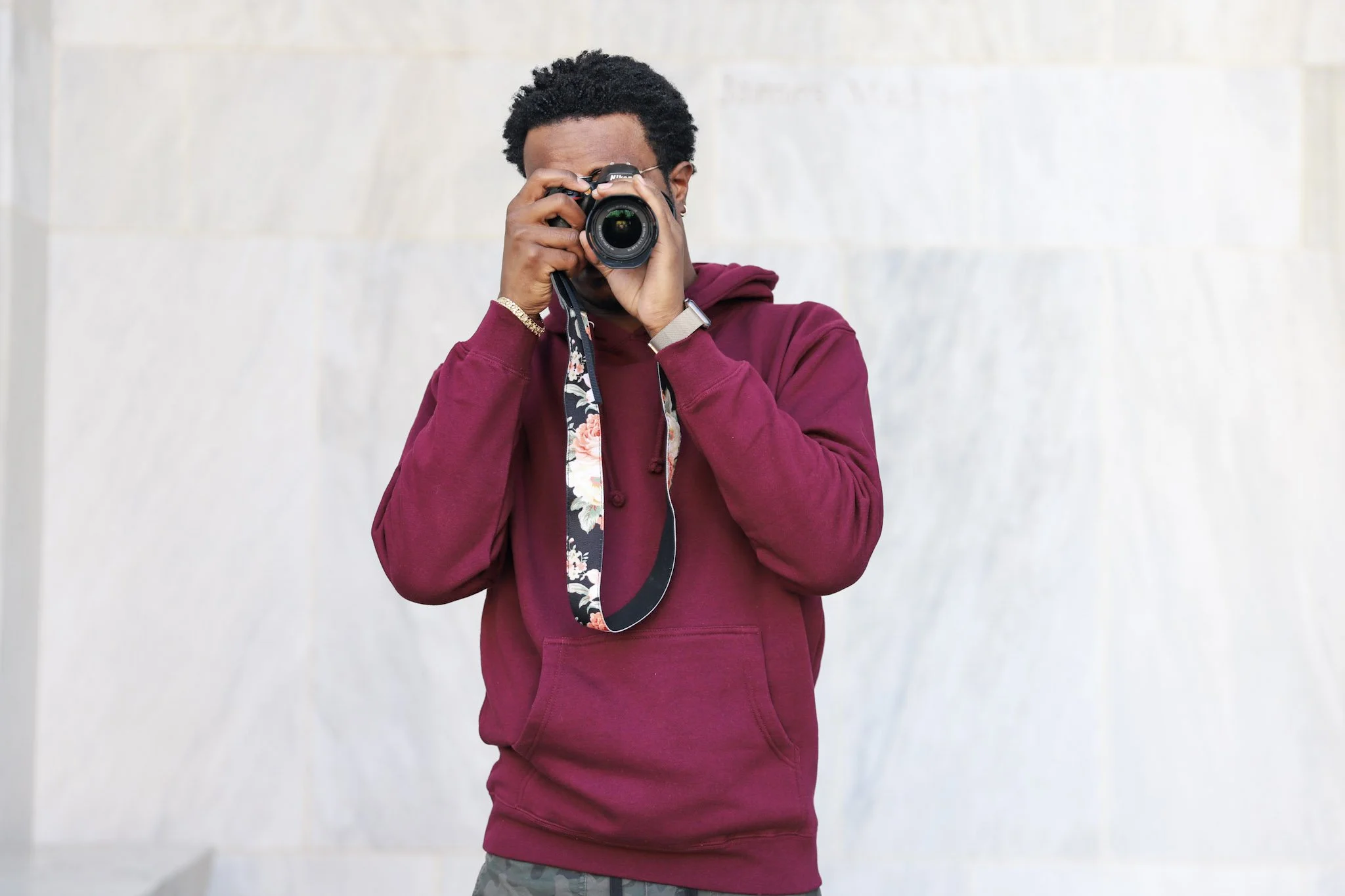 A man with curly hair taking a photo with a camera, wearing a maroon hoodie and standing against a plain light-colored wall.
