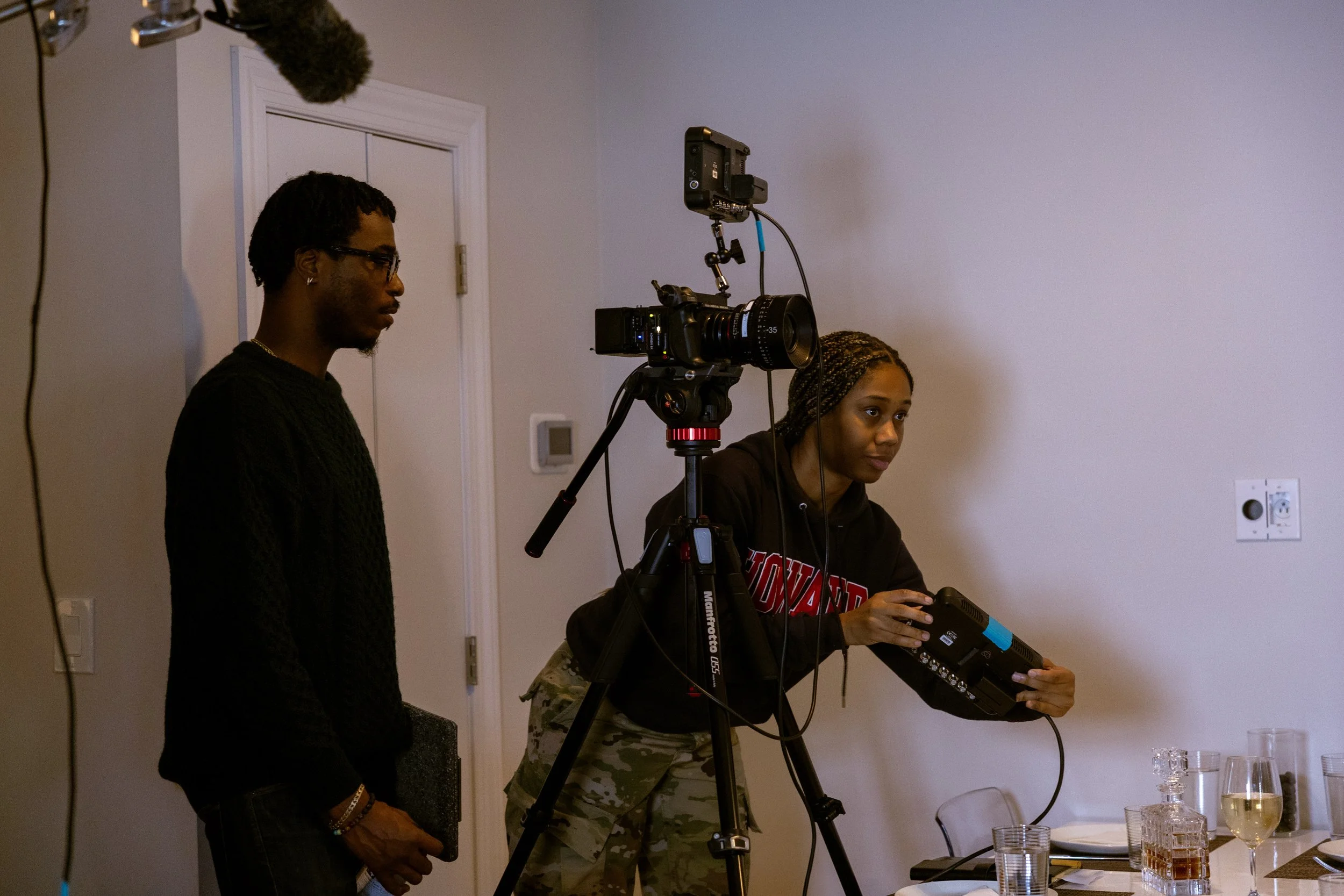 Two people standing beside a camera on a tripod, one woman operating a device, and another person watching, in a room with a table set with glasses and drinks.