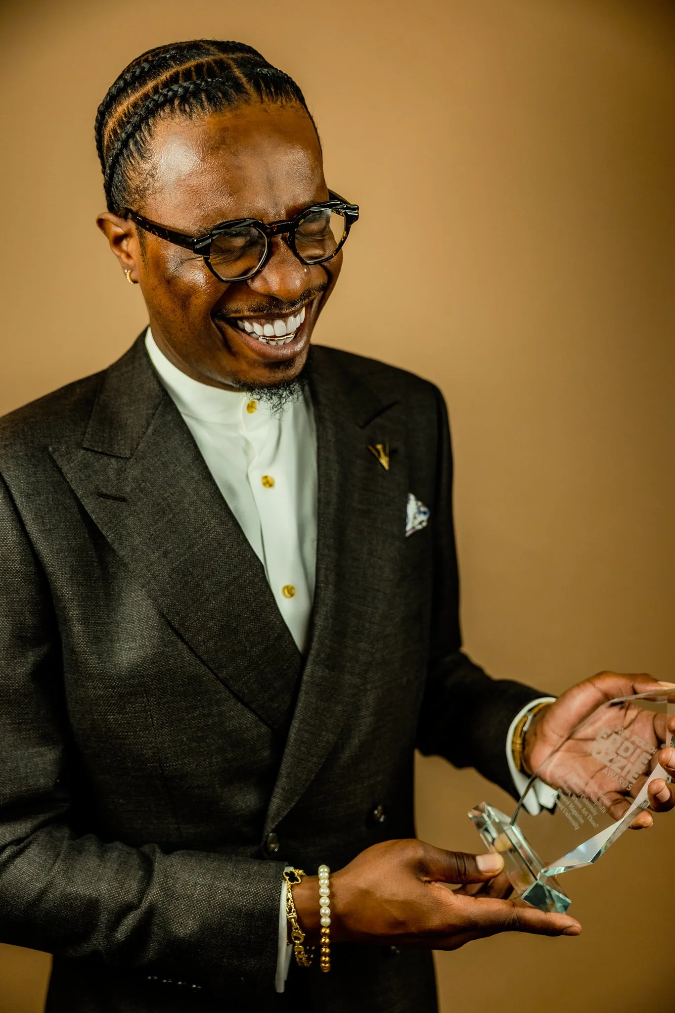A man with cornrow braids and glasses smiling while holding an award and a certificate in a glass stand.