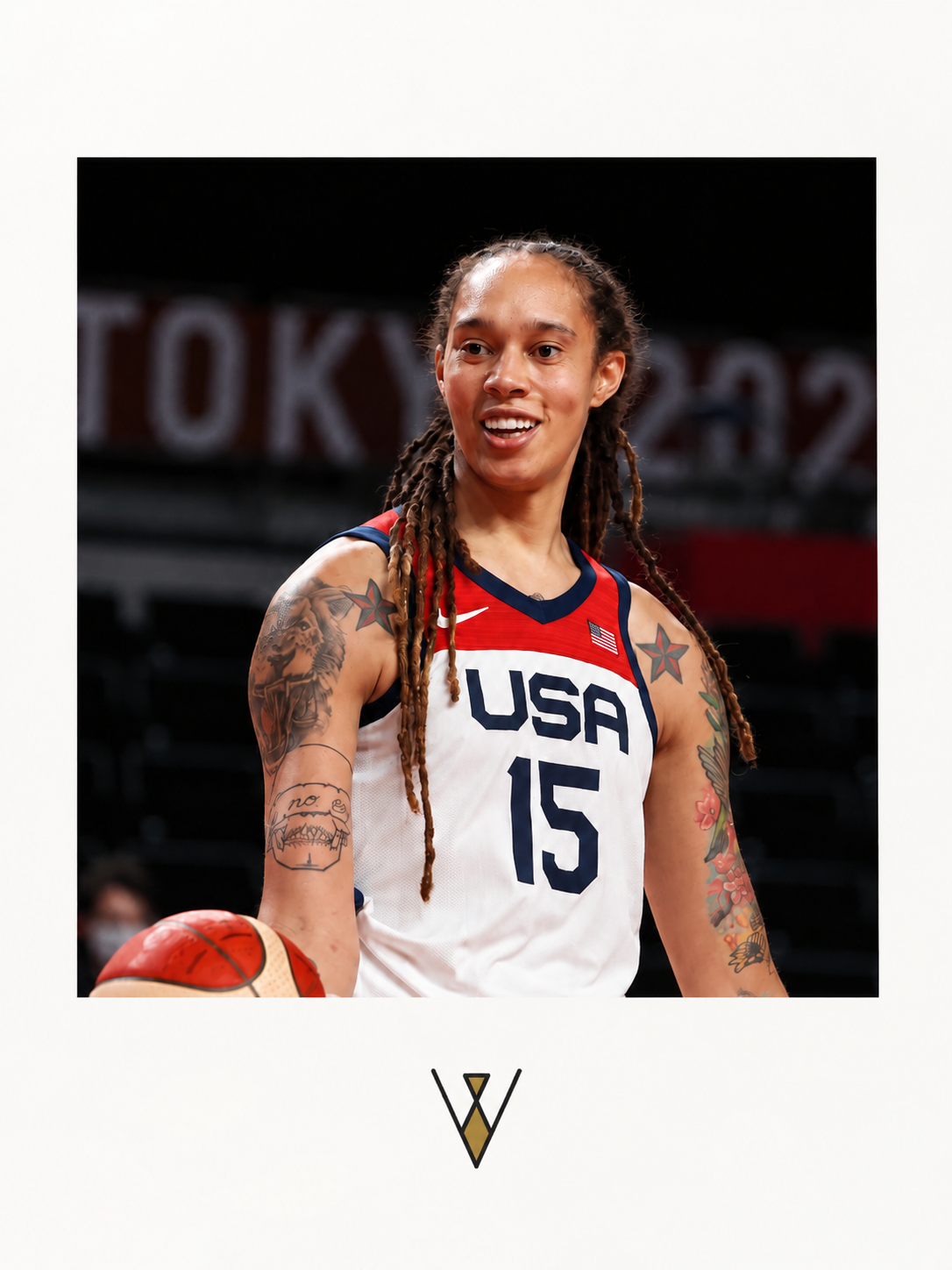 A female basketball player in USA jersey with tattoos, holding a basketball, smiling on the court.