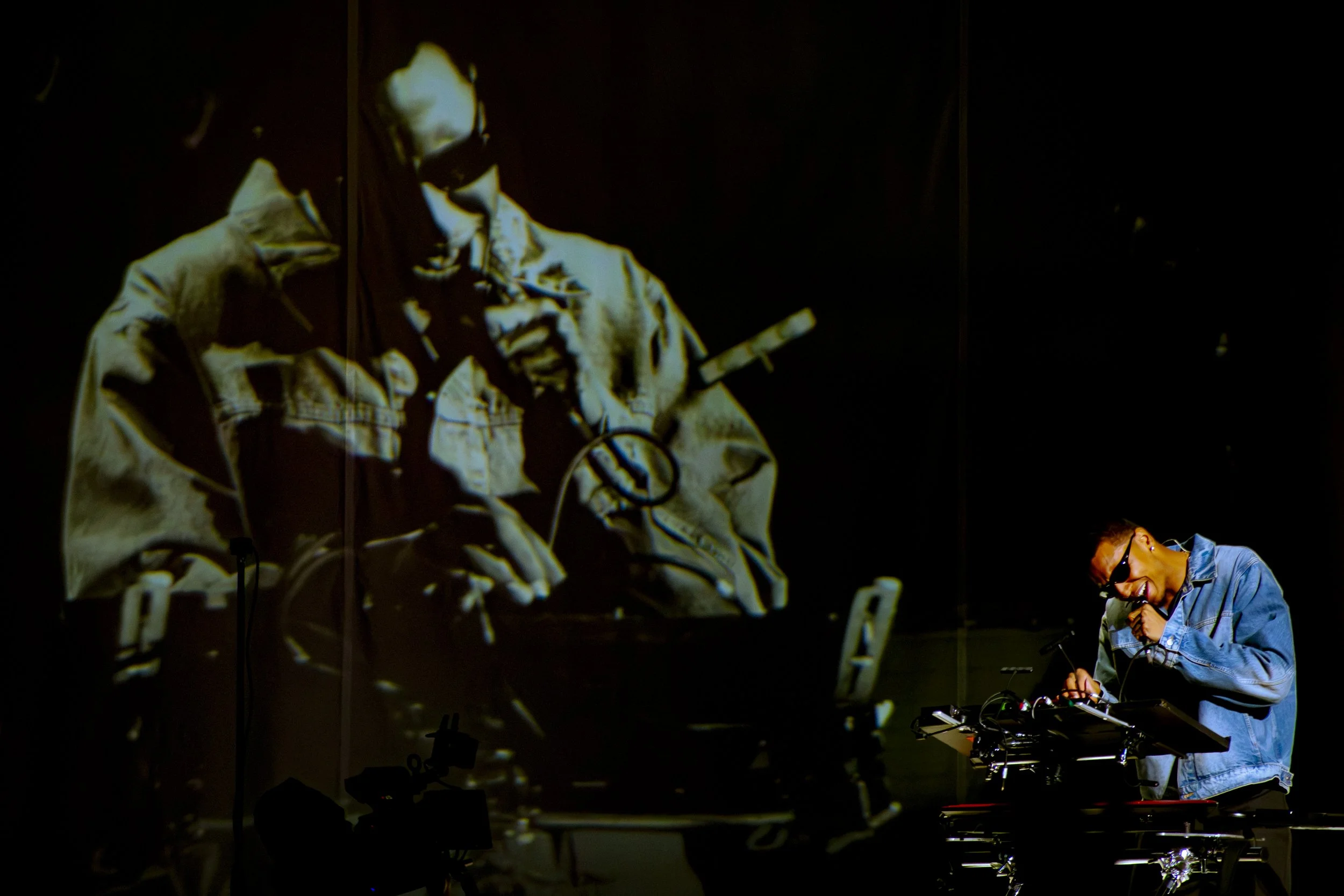 A person with sunglasses wearing a denim jacket performing as a DJ, with a large screen behind them displaying a mirrored image of another person with glasses.