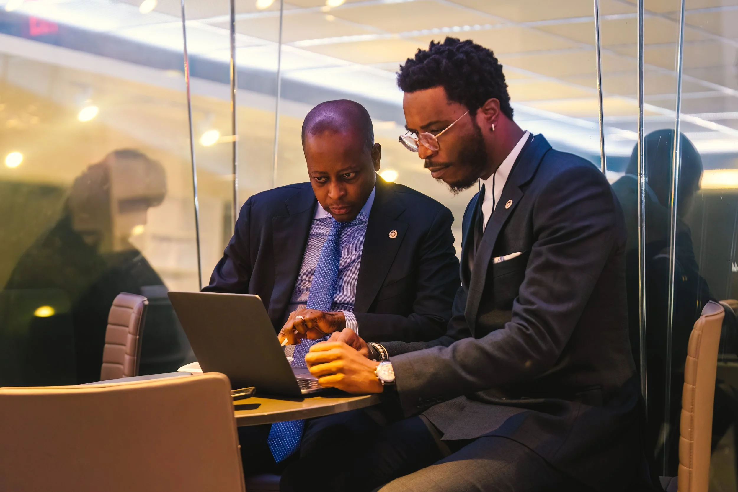 Two men in business suits sitting at a table with a laptop, engaged in a discussion, seen through a glass wall in an office setting.