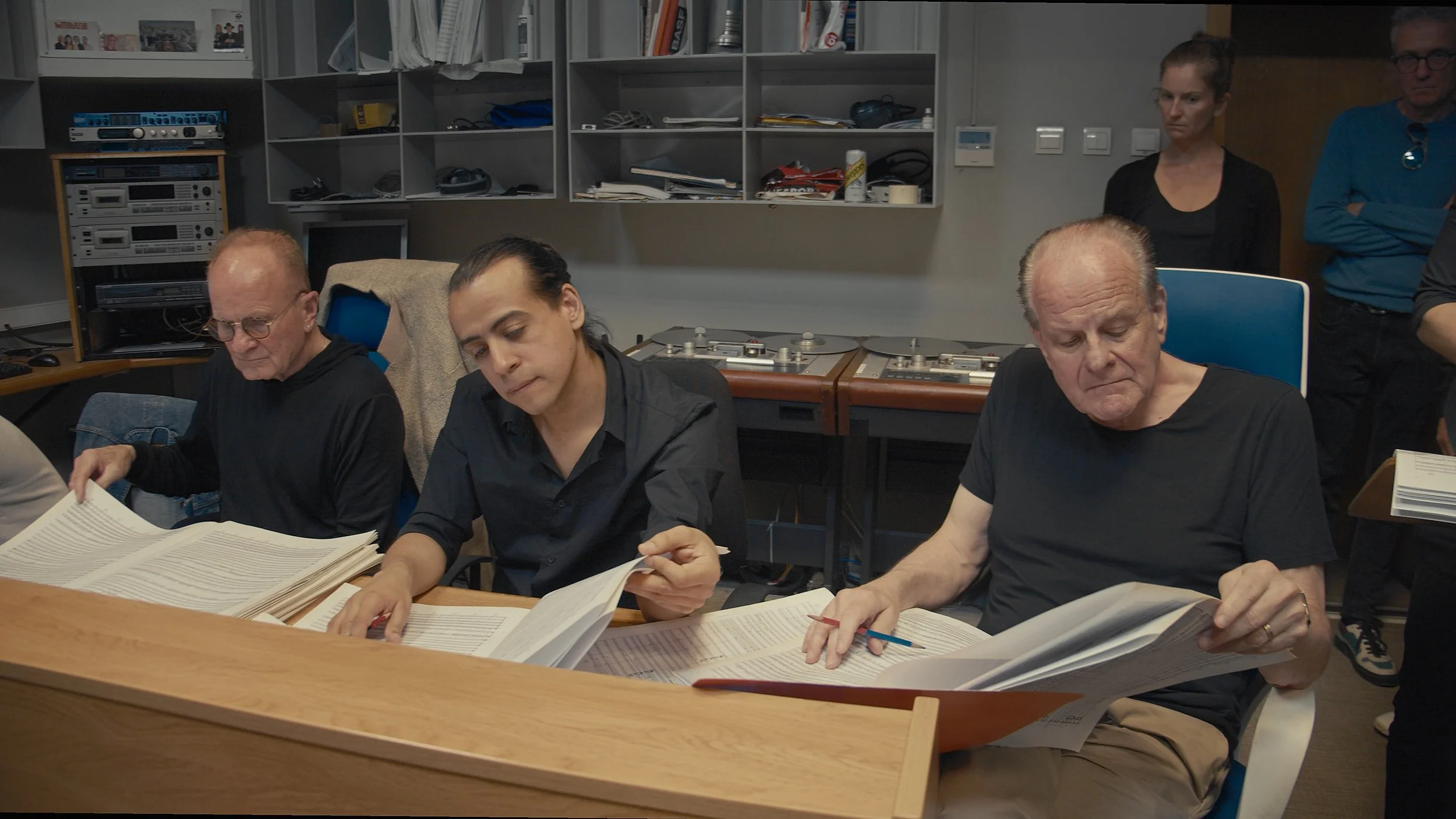 Three men seated at a table reviewing large books, with two women standing behind them in a room with electronic equipment and shelves.