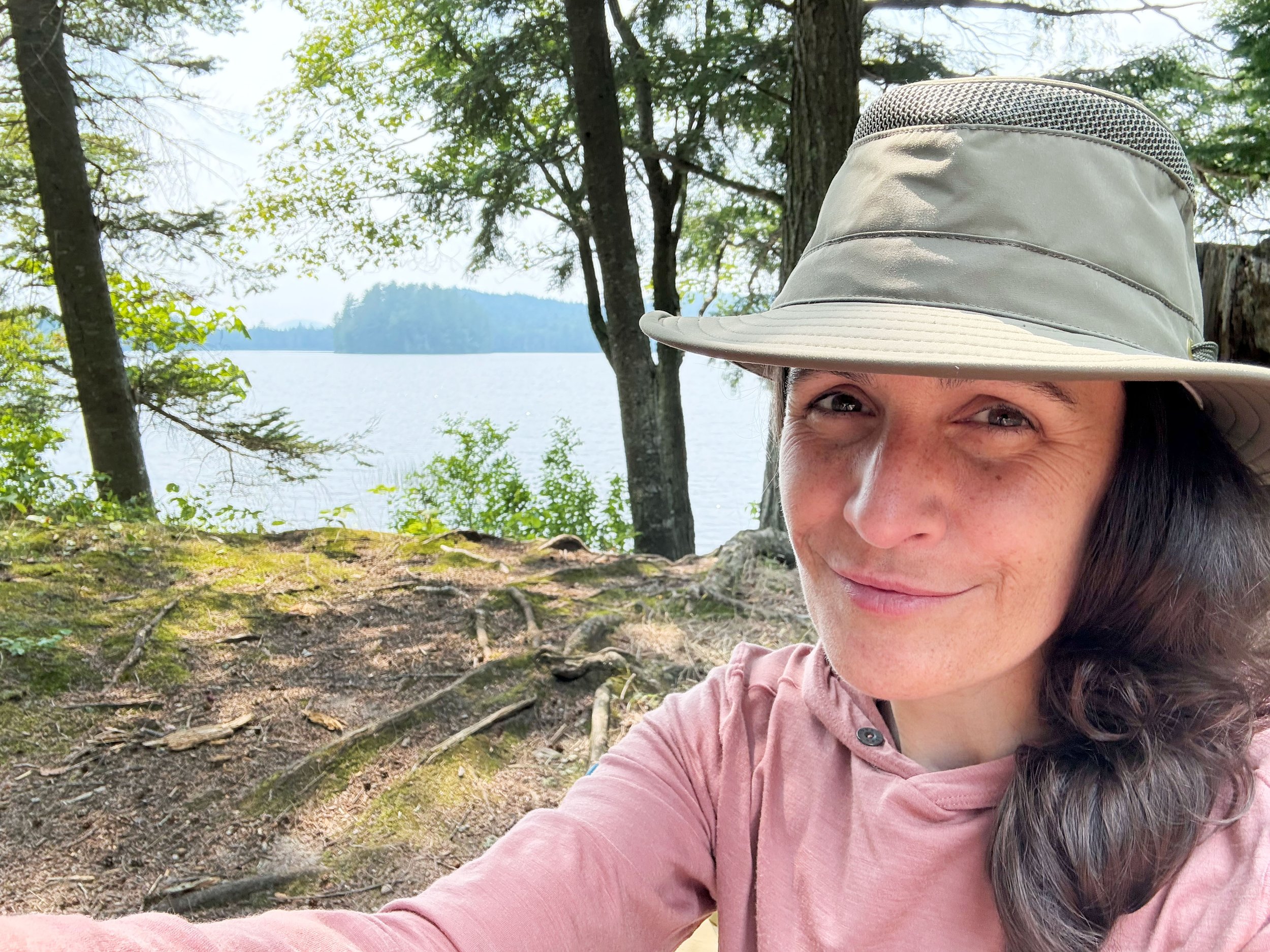 A woman taking a selfie outdoors in a wooded area near a lake, wearing a wide-brimmed hat and a pink long-sleeved shirt, with trees and water in the background.