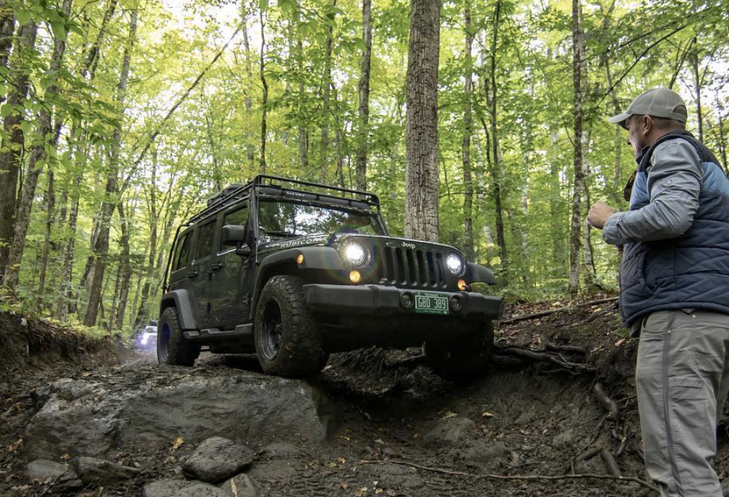A black Jeep Wrangler navigating a rugged, rocky dirt trail in a dense green forest, while a man stands on the side guiding the vehicle over the obstacles.