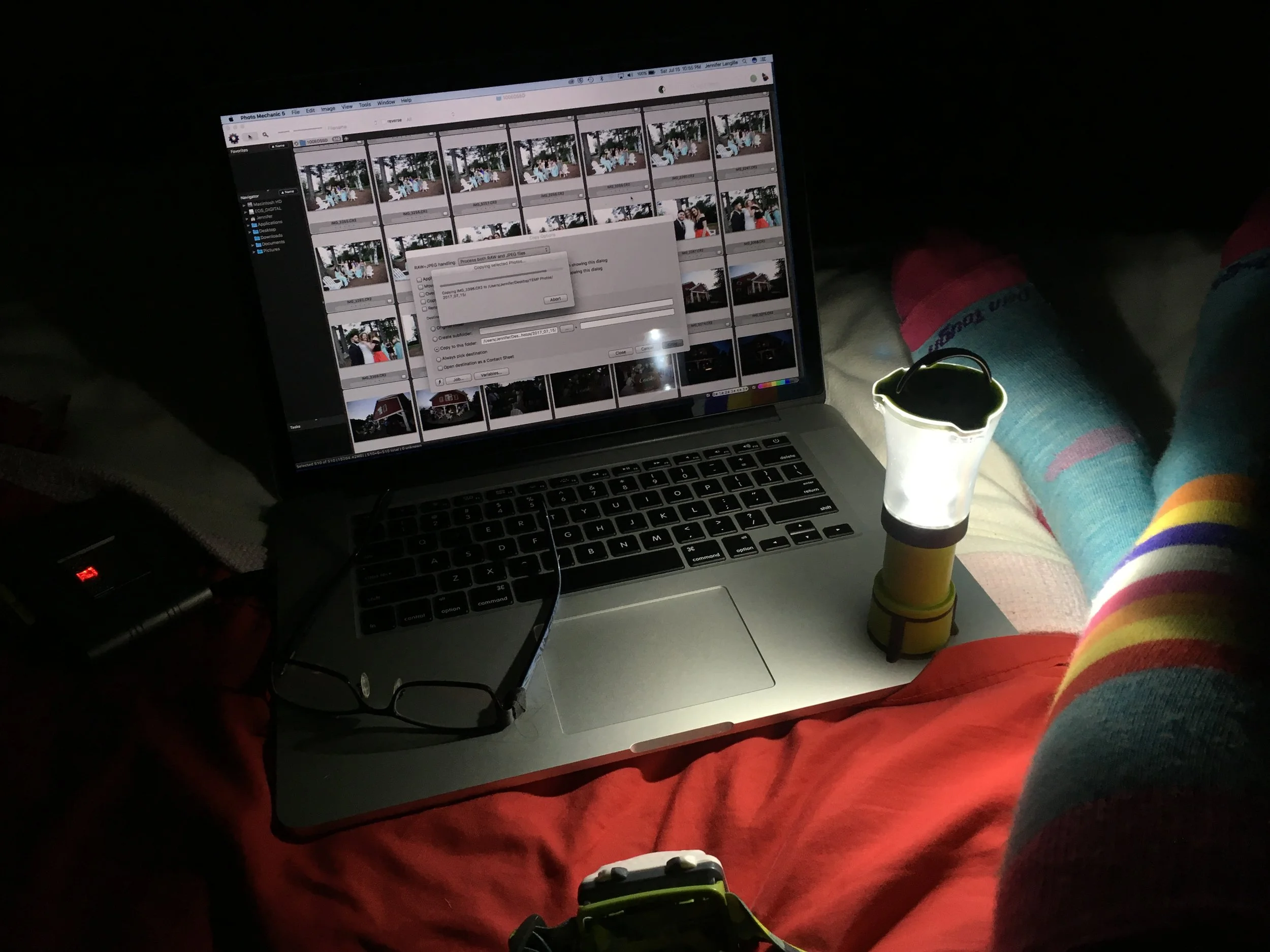 first-person view of working inside a dark tent or room, showing an open laptop displaying photo editing software alongside a small lit camping lantern, eyeglasses, and legs wearing bright, colorful socks.