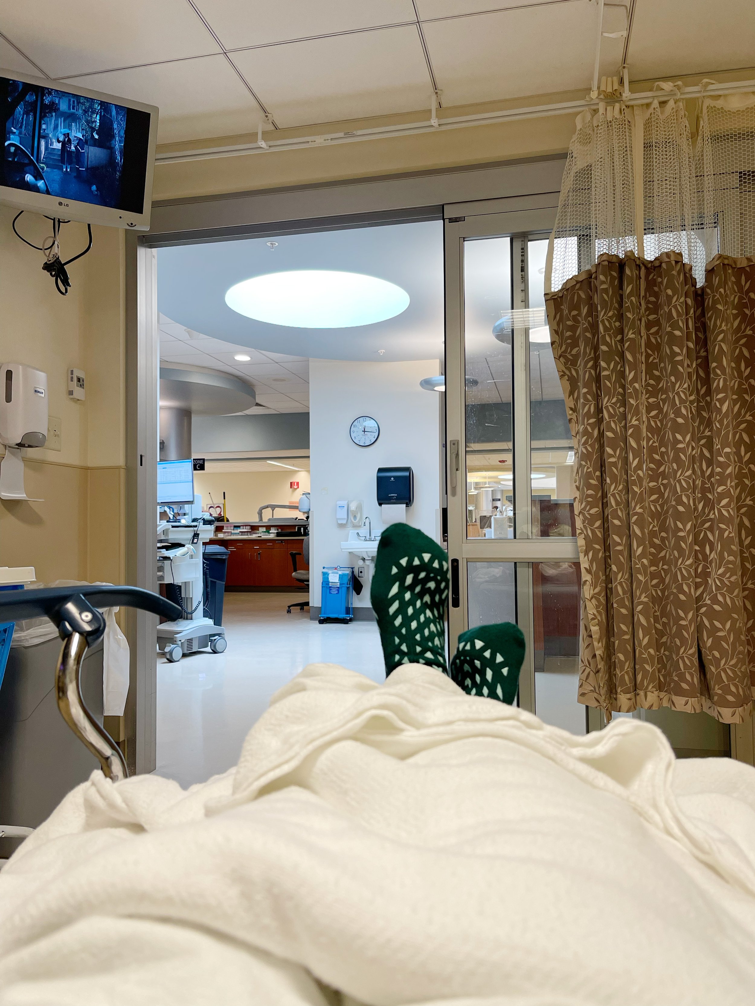 A first-person perspective from a hospital bed, showing a patient's feet in green non-slip hospital socks propped up under a white blanket, looking out an open sliding glass door toward a brightly lit nurse's station.