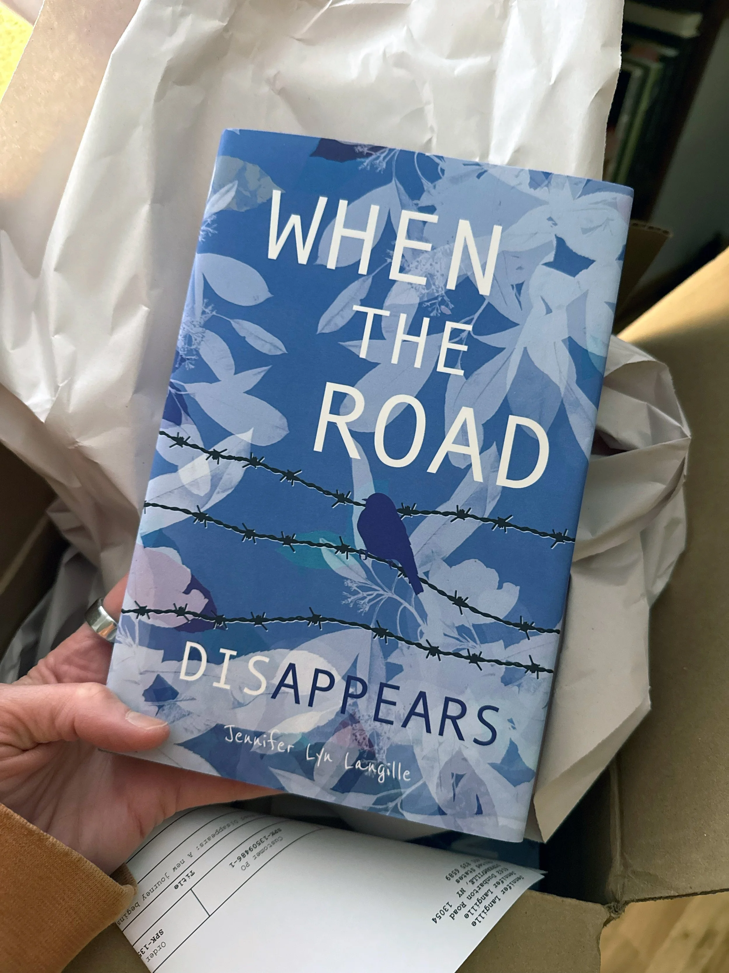 Book titled 'When the Road Disappears' by Jennifer Lyn Langille, held in hand with a background of a paper bag and some packaging or receipts.