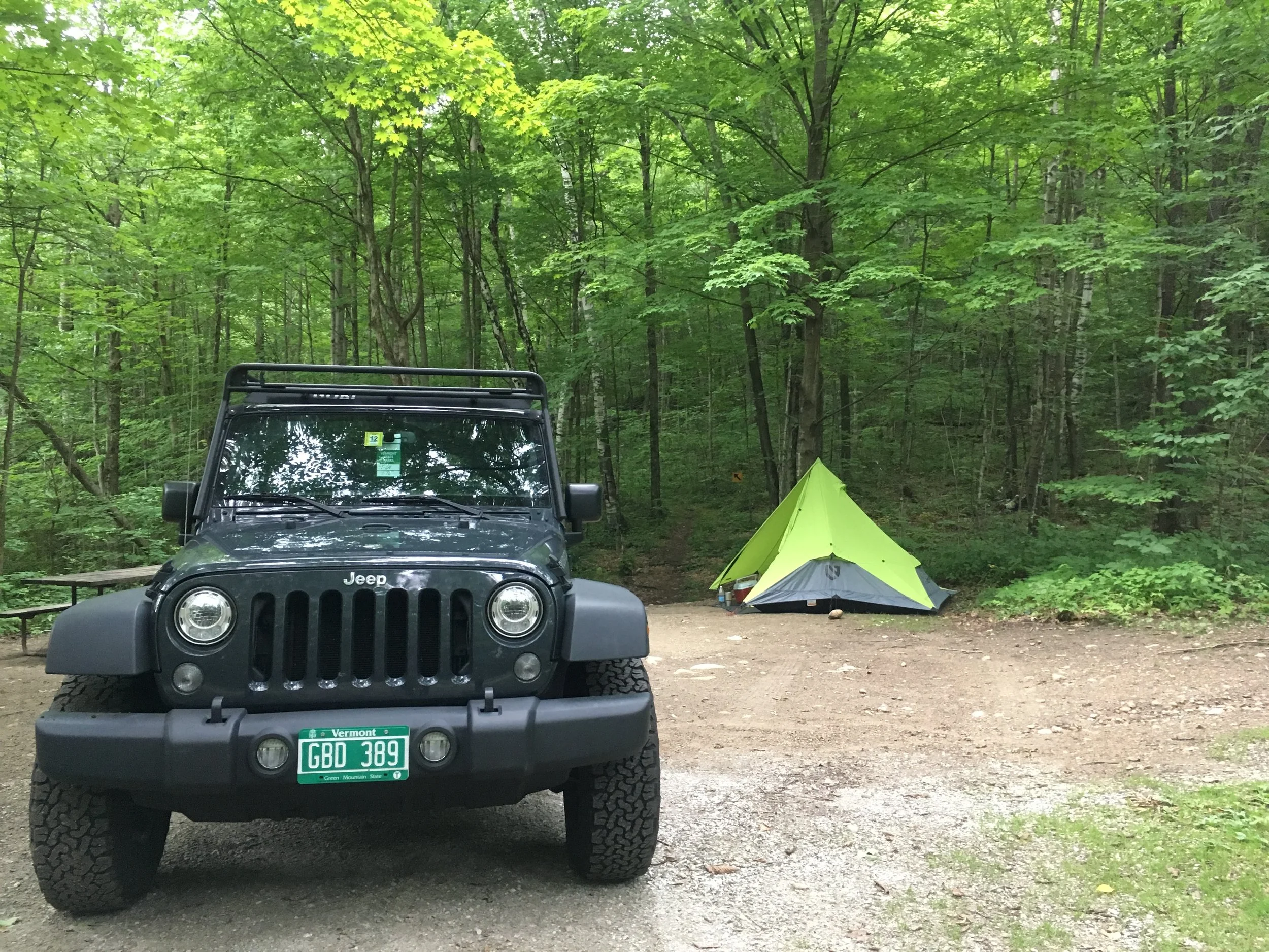 A black Jeep Wrangler parked at a wooded campsite next to a bright green pyramid-shaped tent tucked among tall trees.
