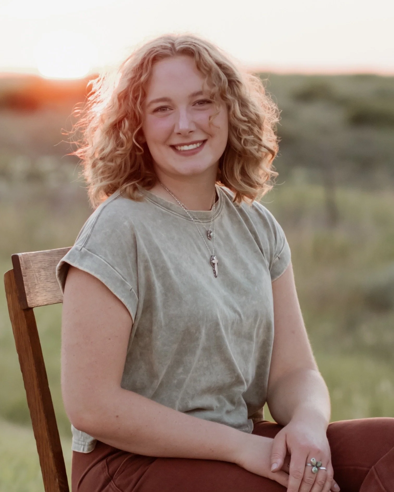 A young woman with curly blonde hair smiling outdoors during sunset, sitting on a wooden chair, wearing a green t-shirt, brown pants, and handmade sterling silver jewelry.
