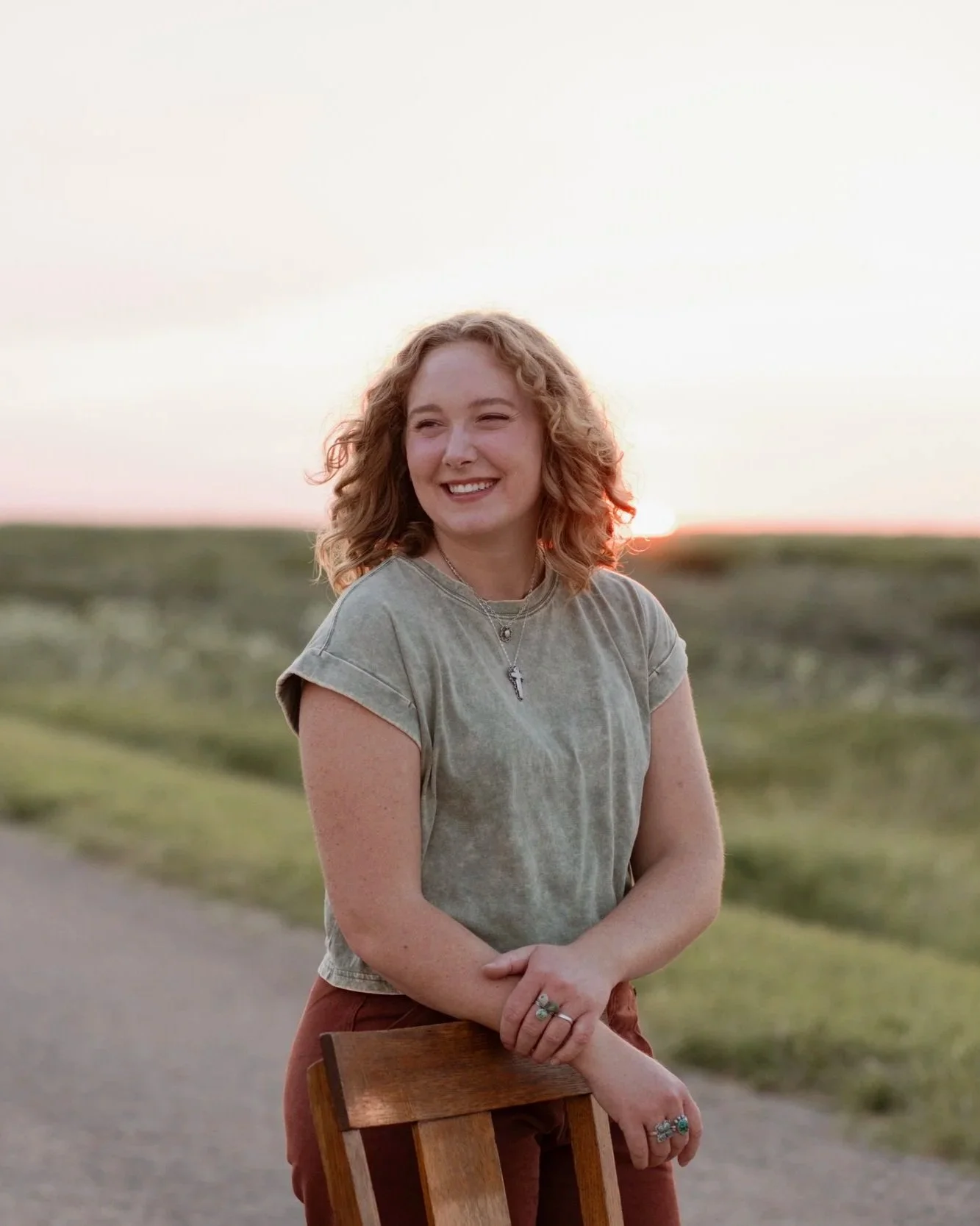 A smiling young woman with curly blonde hair, wearing a green t-shirt and handmade sterling silver jewelry, standing outdoors during sunset with a landscape of green fields in the background.