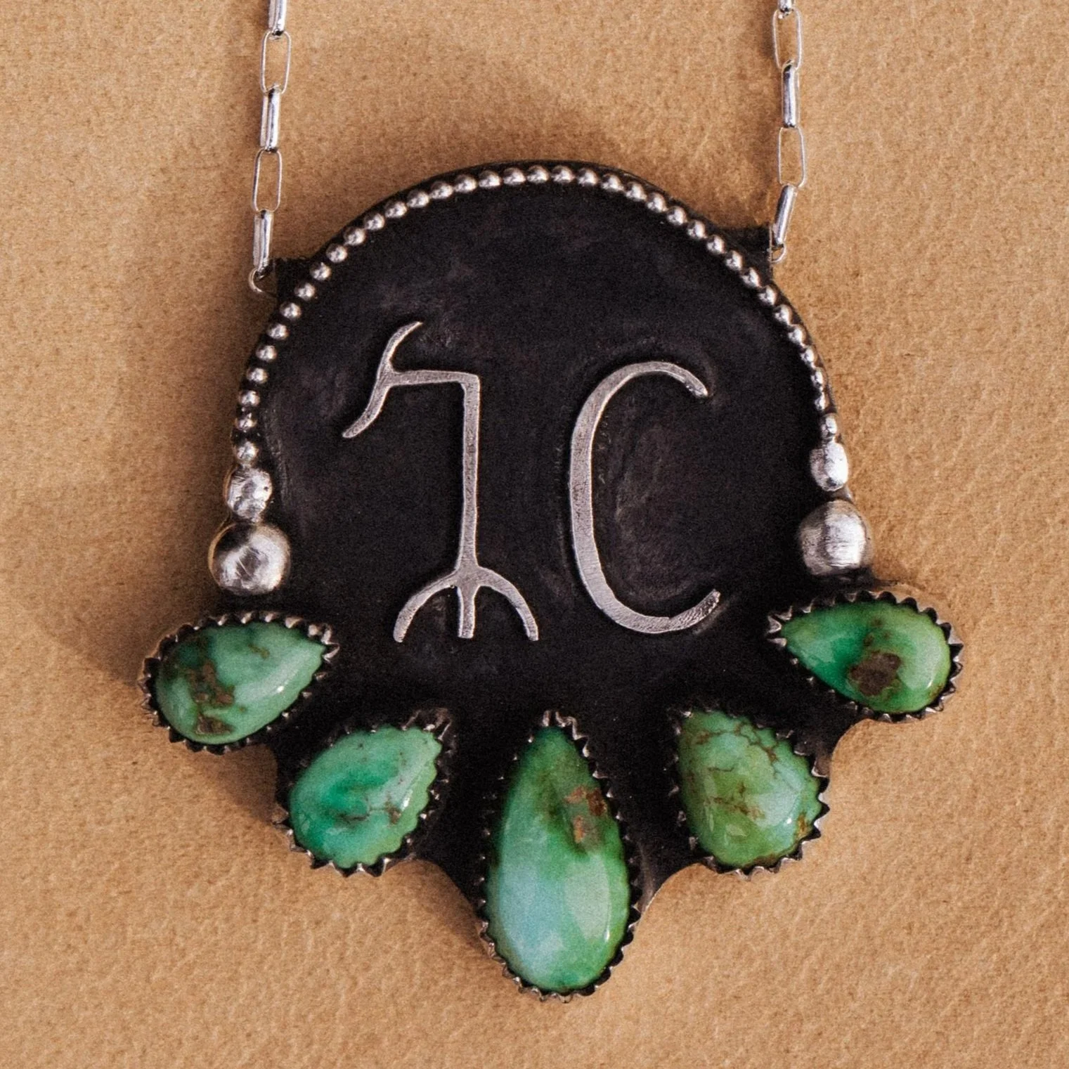 Necklace with a black background featuring silver cattle brand markings surrounded by green turquoise stones.