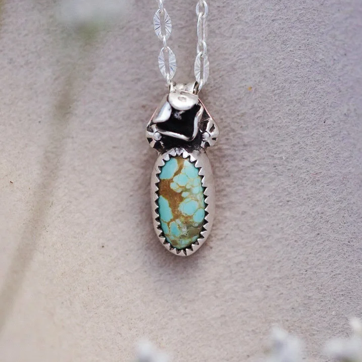 Sterling silver necklace with oval turquoise pendant and flower-shaped accent on top, hanging against a neutral background.