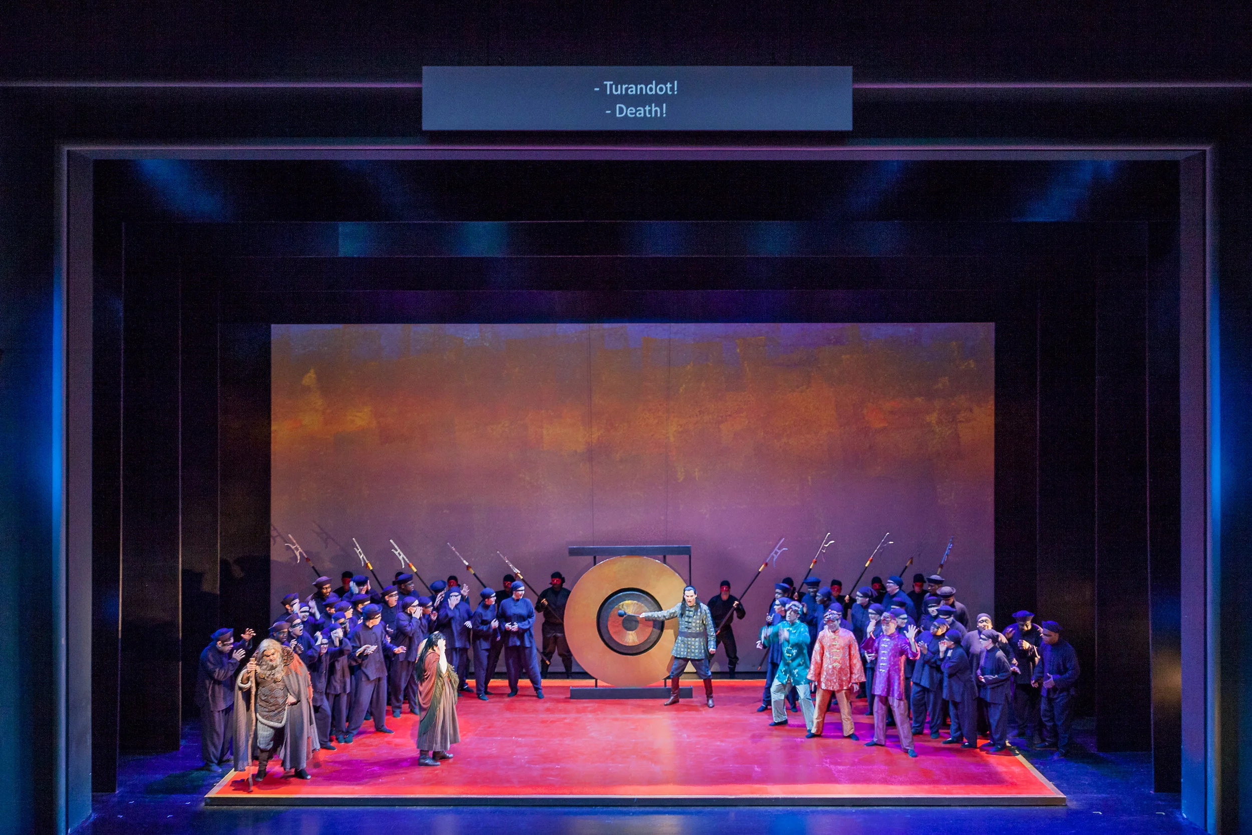 Prince Calaf, Derek Taylor, rings the gong accepting the challenge, while his father Timur, Ricardo Lugo, and the slave girl Liu, Danielle Pastin, watch in fear; Ping, Pang, and Pang also seen on the right side. Photo by Ben Schill Photography; courtesy of Virginia Opera.