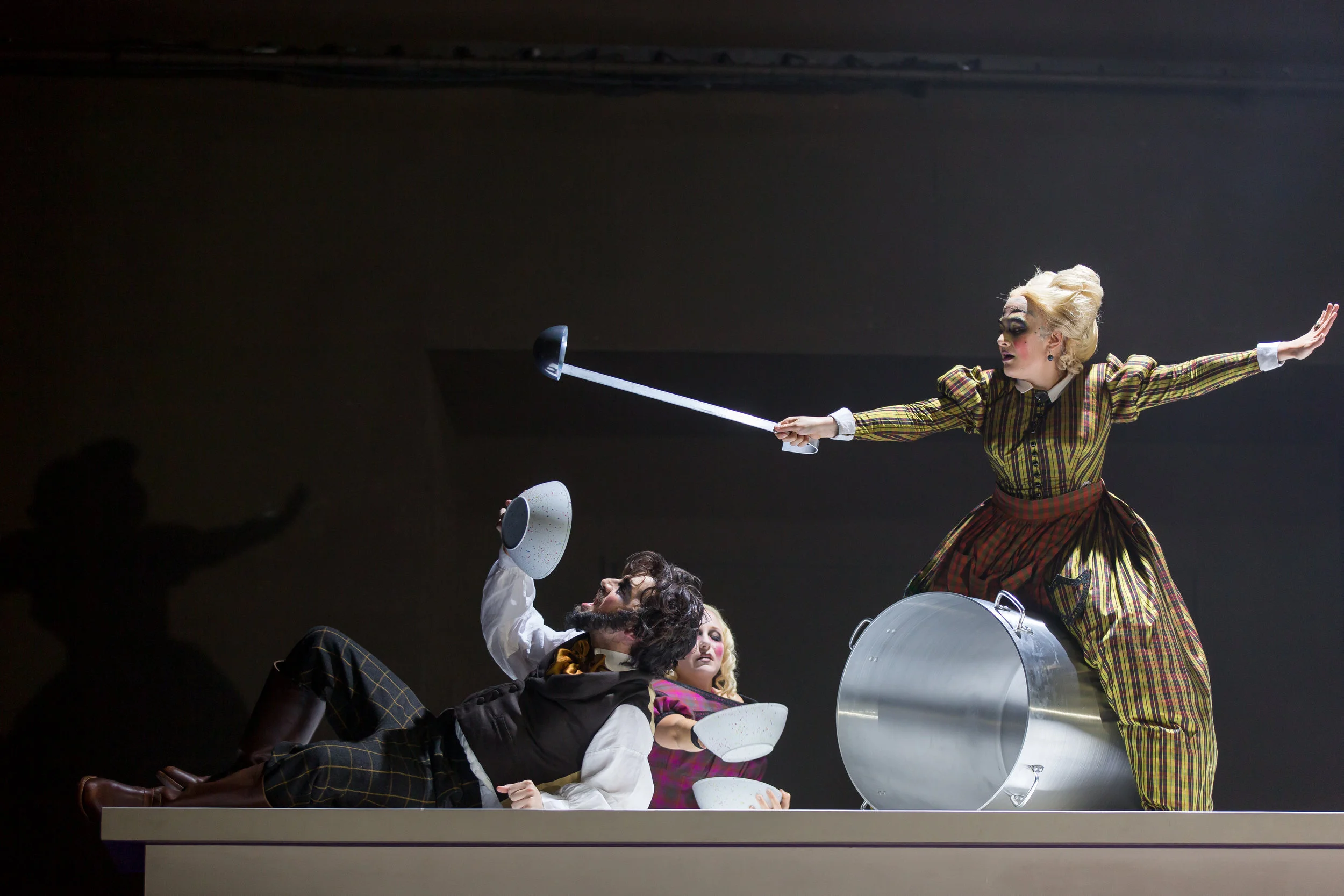 From The Juniper Tree : Ben Edquist, the father, is fed a stew containing his son; Madison Leonard, the daugher, assists; and Annie Rosen, the stepmother serves the stew atop a the large table; photo by Scott Suchman and courtesy of Wolf Trap Opera.