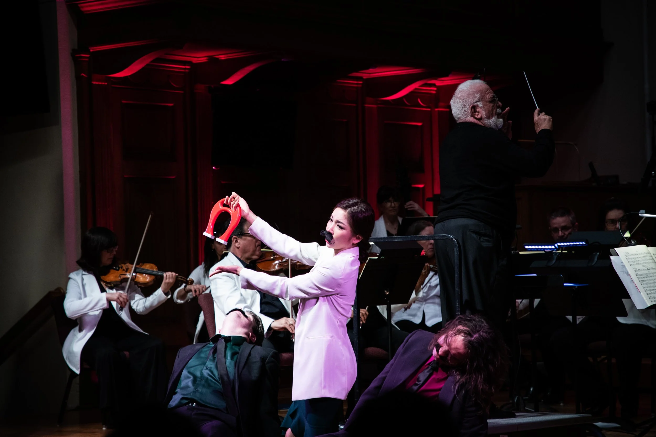 A comedic moment of Cosi fan tutte ; soprano Nanyoung Song as Despina in disguise holds the magic cure for the passed out boys and, Conductor Louis Salemno holds the baton in the background. Photo by Sam Trotman; courtesy of Maryland Lyric Opera.
