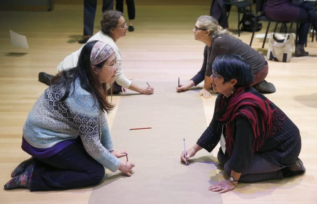 Women sitting on a wooden floor, engaged in drawing or writing on large sheets of paper laid out in front of them.